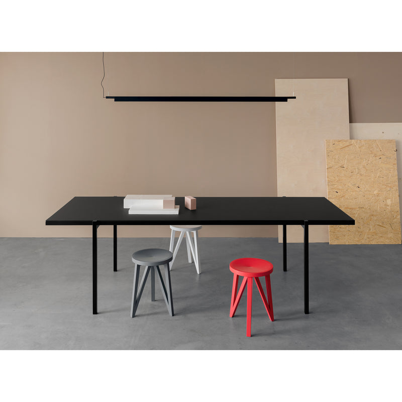The minimalist room showcases a sleek DL5 Neo Table by Loehr, paired with three stools in gray, white, and red, reflecting the De Stijl style. A long black light fixture hangs above while leaning plywood boards rest against a beige wall. The gray concrete floor enhances the contemporary feel.