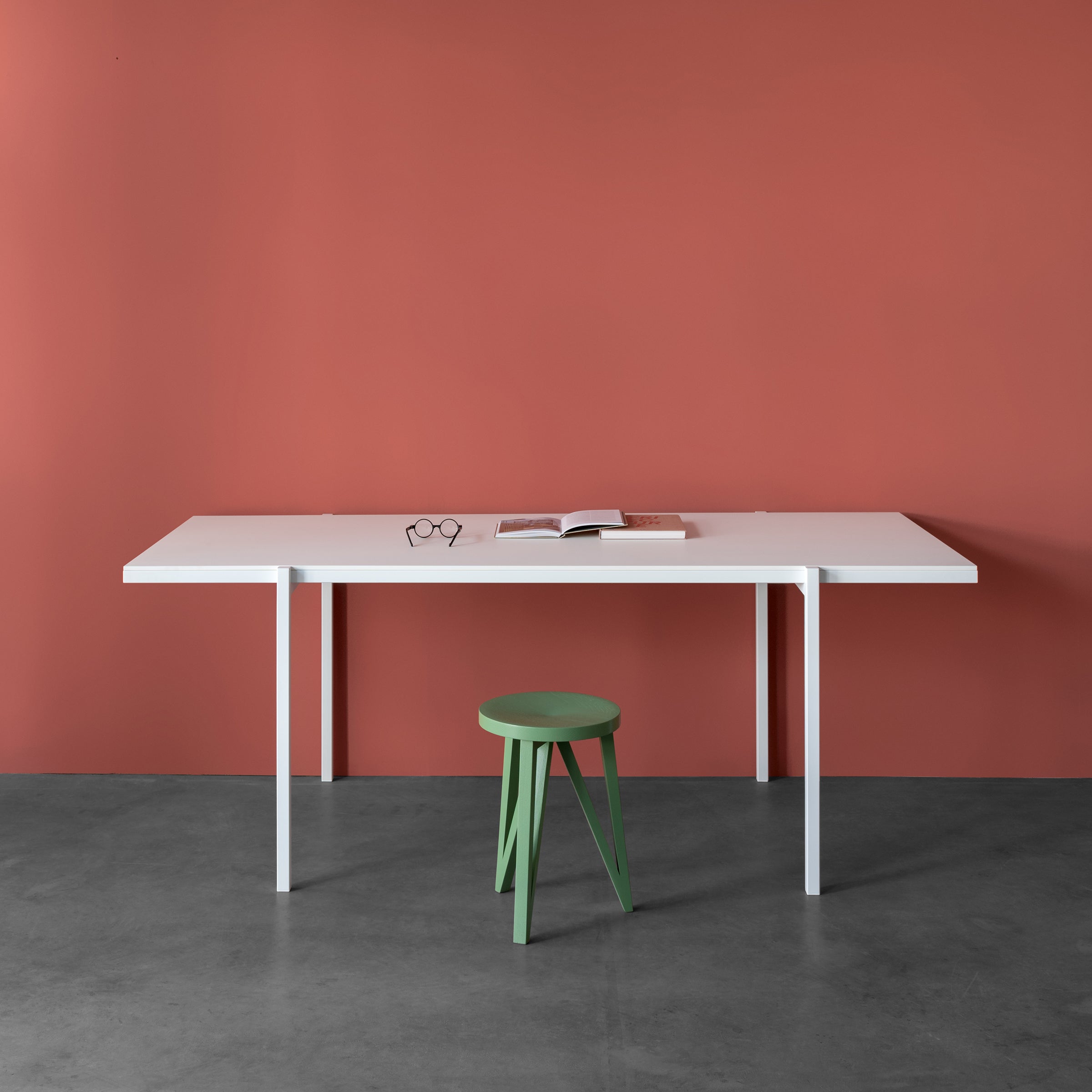 A minimalist scene showcases a Loehr DL5 Neo Table with steel tubes against a red wall. An open book and glasses sit on the table, while a green stool, reminiscent of De Stijl aesthetics, stands in front. The floor is smooth gray concrete.