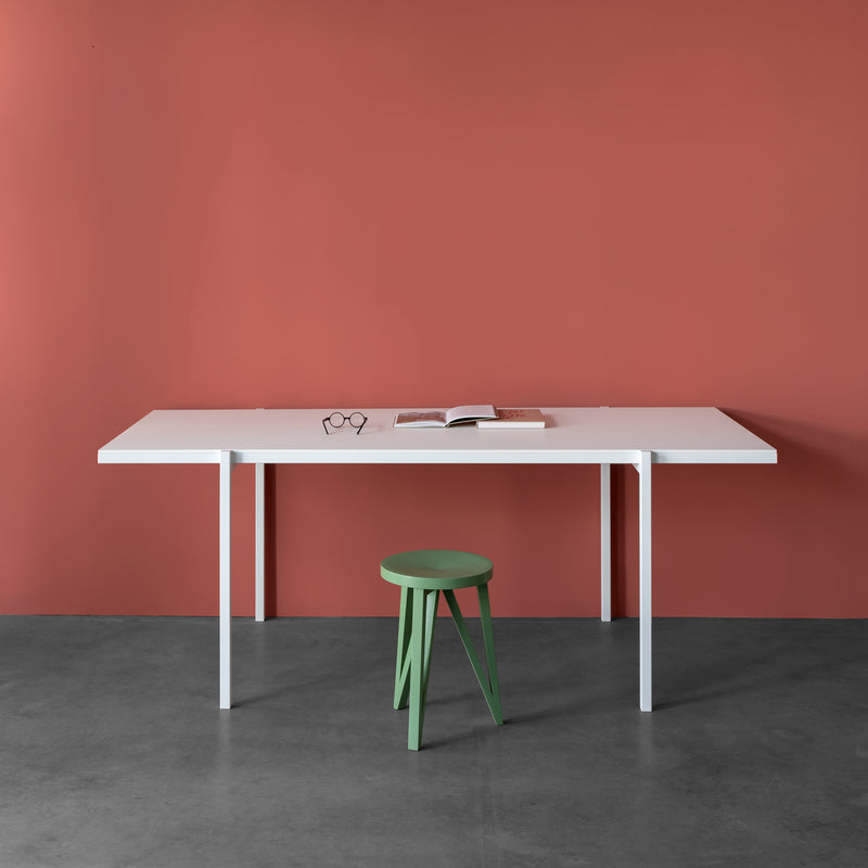 A minimalist scene showcases a Loehr DL5 Neo Table with steel tubes against a red wall. An open book and glasses sit on the table, while a green stool, reminiscent of De Stijl aesthetics, stands in front. The floor is smooth gray concrete.