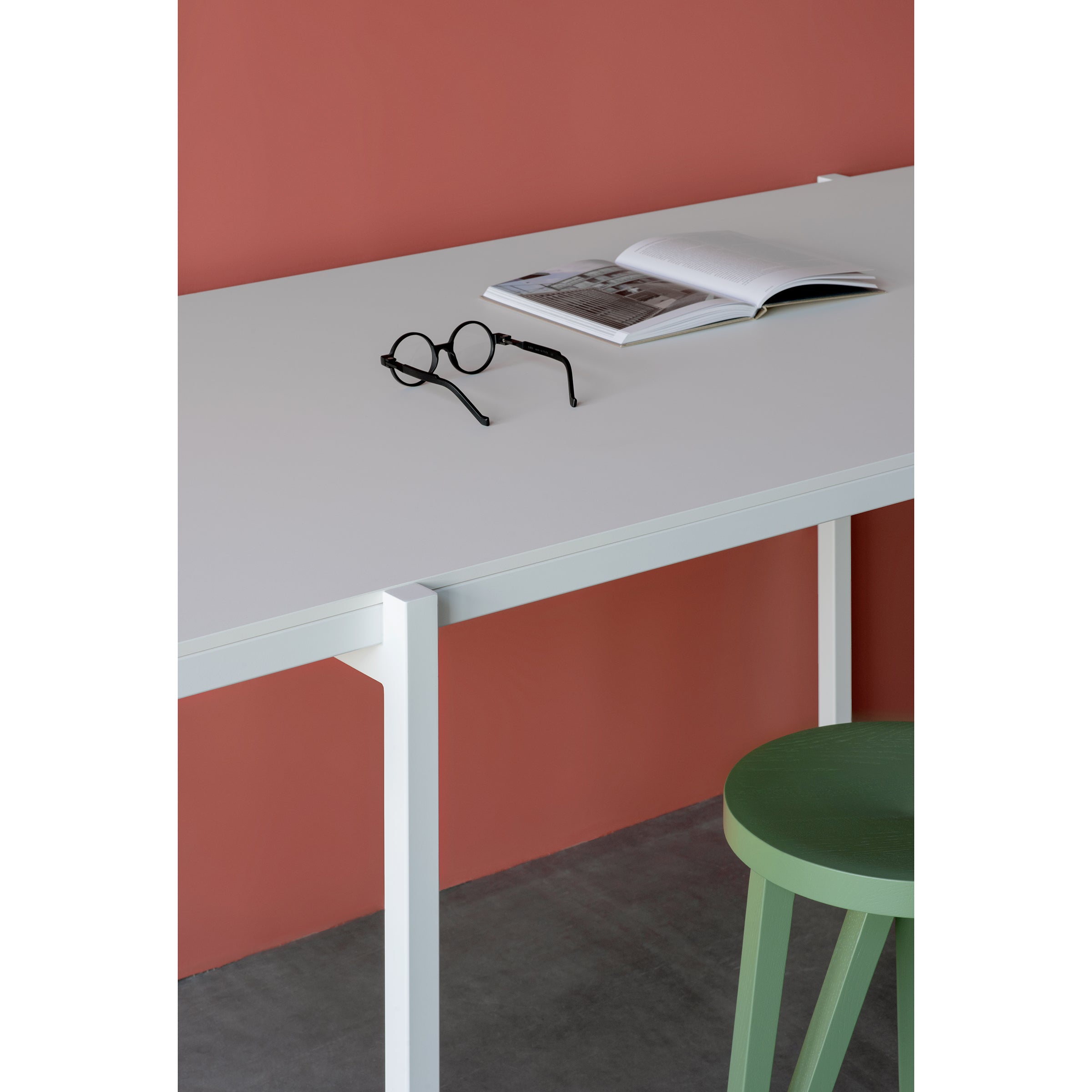 A DL5 Neo Table by Loehr in white is set against a red wall, featuring an open book and round glasses on top. A De Stijl-inspired green stool is partially visible in front of the table on a gray floor.