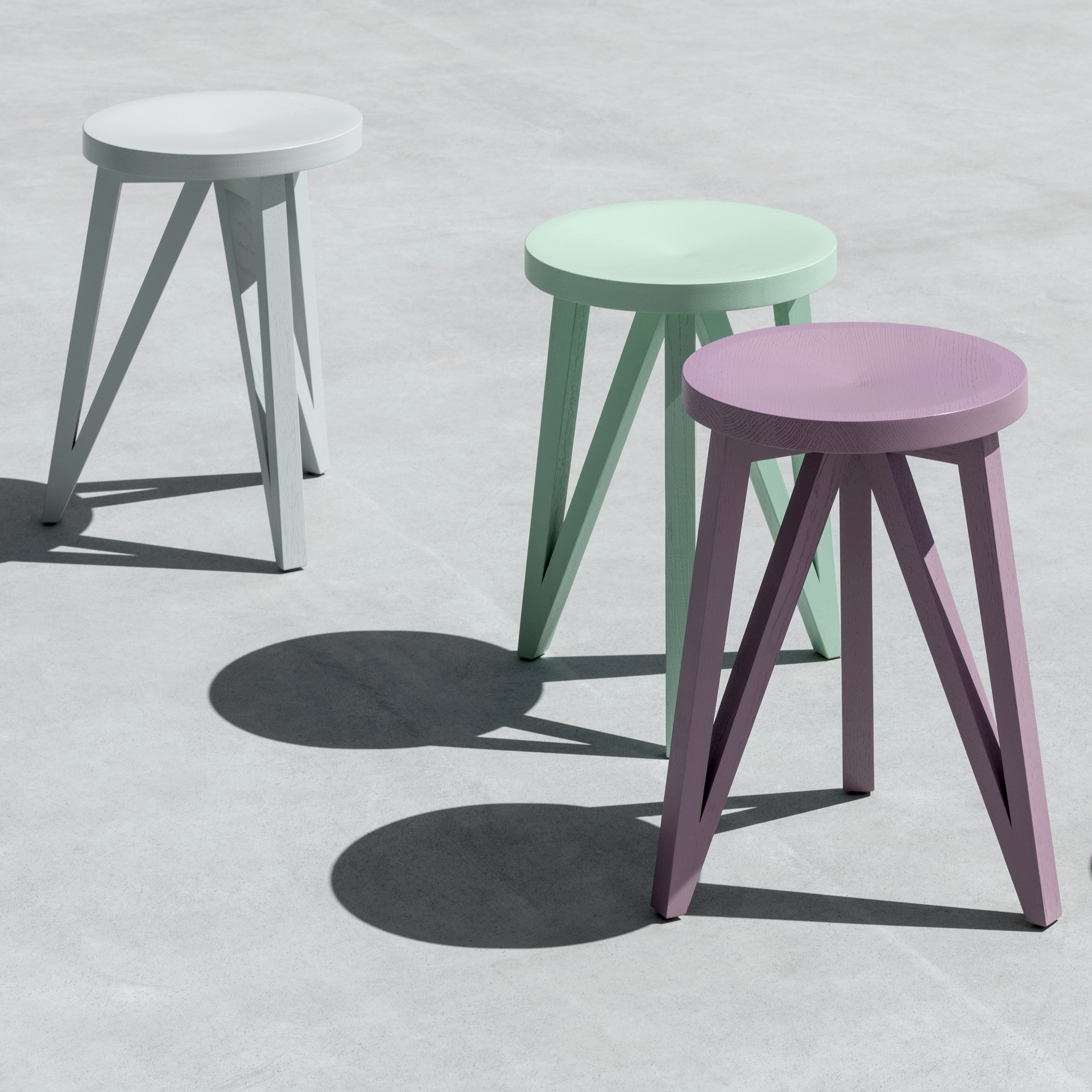 Three JL1 Faber Stools from Loehr lie on a concrete surface in pastel shades of white, mint, and lavender. Their geometric design casts distinct shadows, highlighting the minimalist elegance synonymous with northern European style.