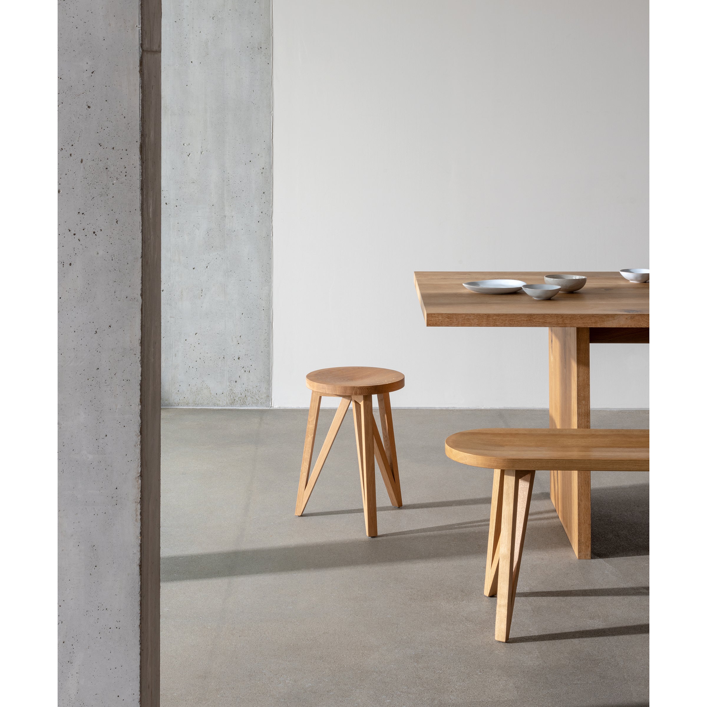 The minimalist dining area highlights northern European design elegance with solid oak furniture—a wooden bench, table, and a JL1 Faber Stool by Loehr—on a sleek concrete floor. Textured concrete pillars and light-colored walls complement artfully arranged FABER collection bowls on the table.