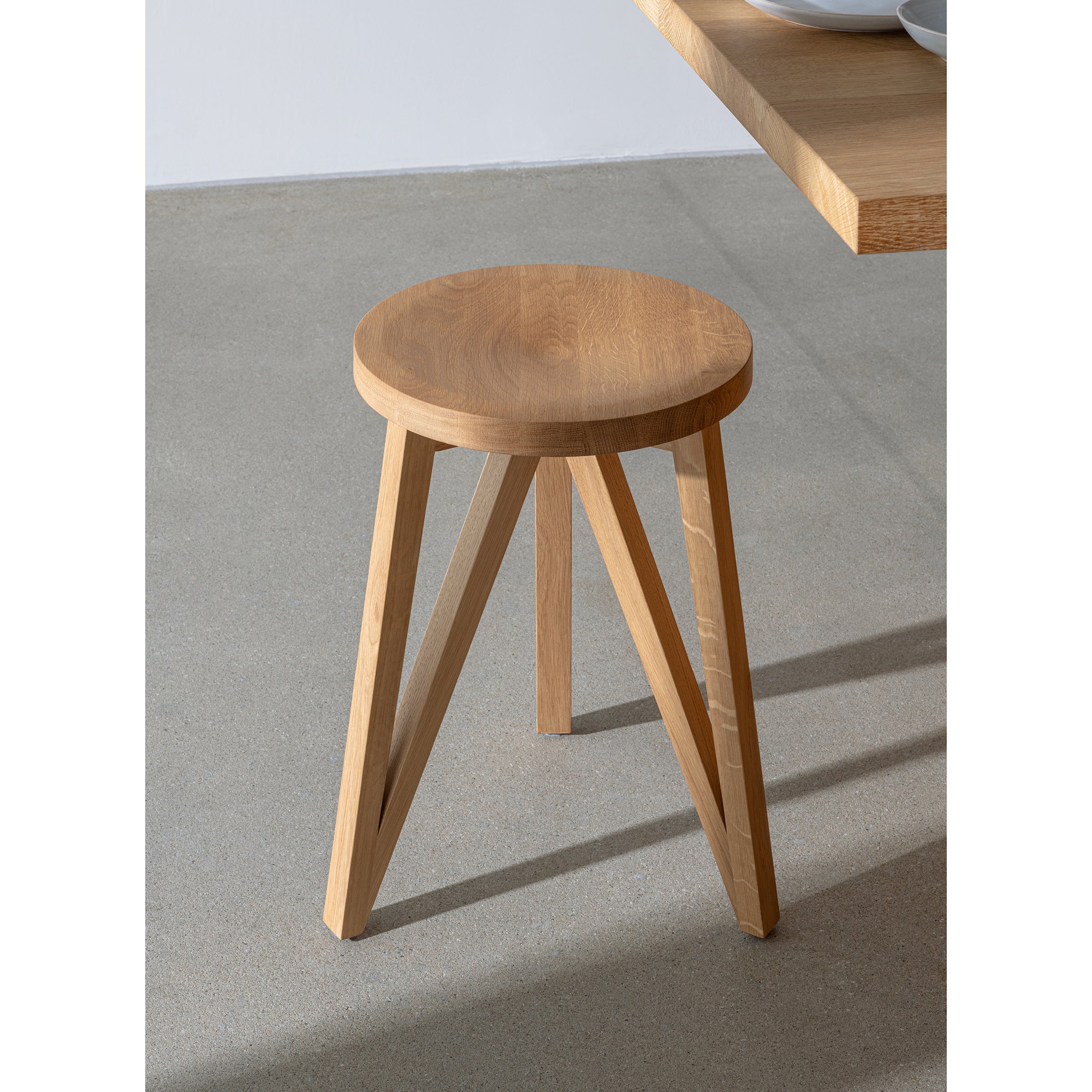 The JL1 Faber Stool by Loehr, featuring a round seat and four straight legs in northern European design, rests on a light gray concrete floor. A wooden table from the FABER collection nearby casts a shadow, enhancing the settings minimalistic and modern appeal.