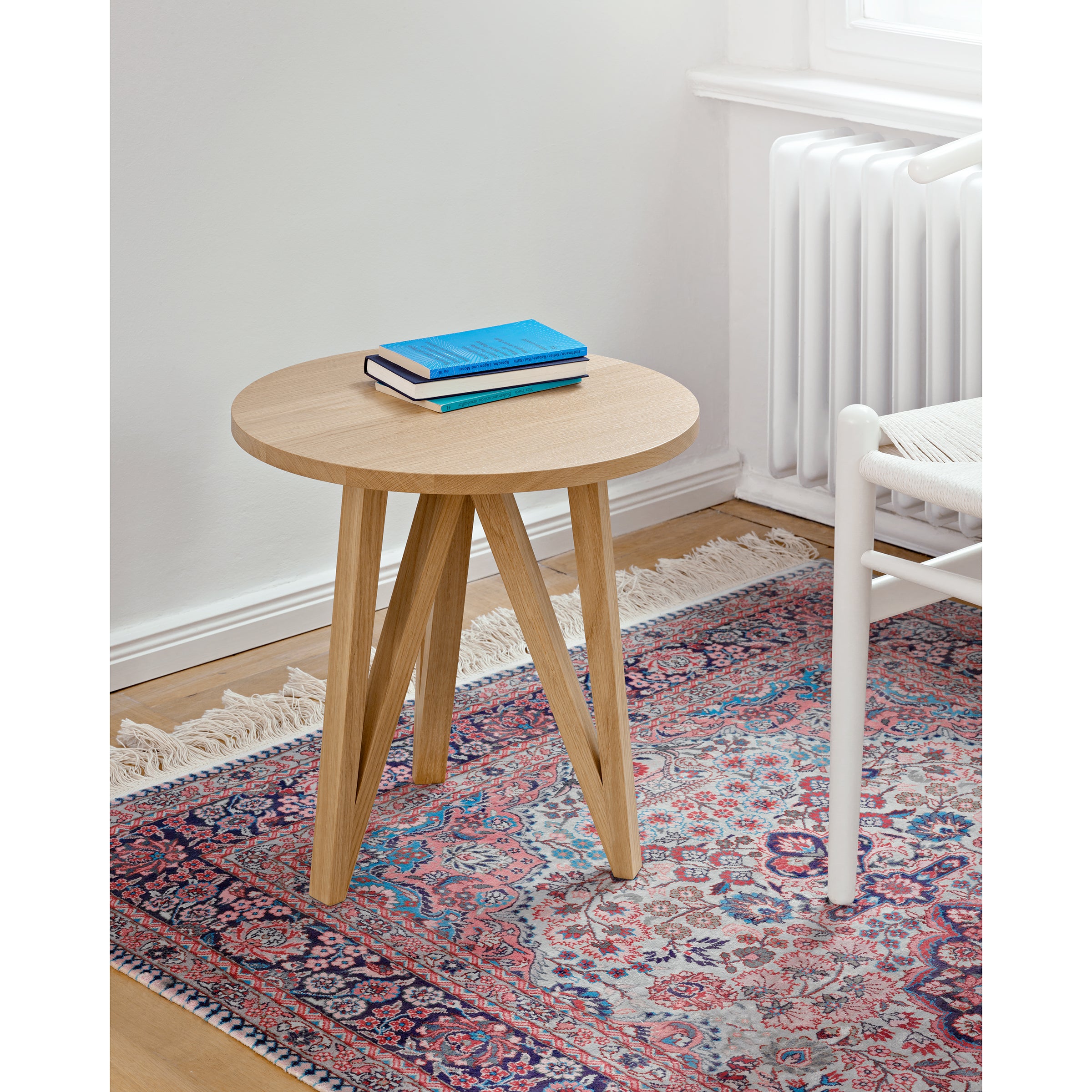 The JL2 Faber Side Table by Loehr, crafted from solid oak with a unique silhouette and angled legs, holds two stacked books, one blue and one pink. It sits on a colorful patterned rug in a bright room next to a white radiator and a woven-seat white chair.