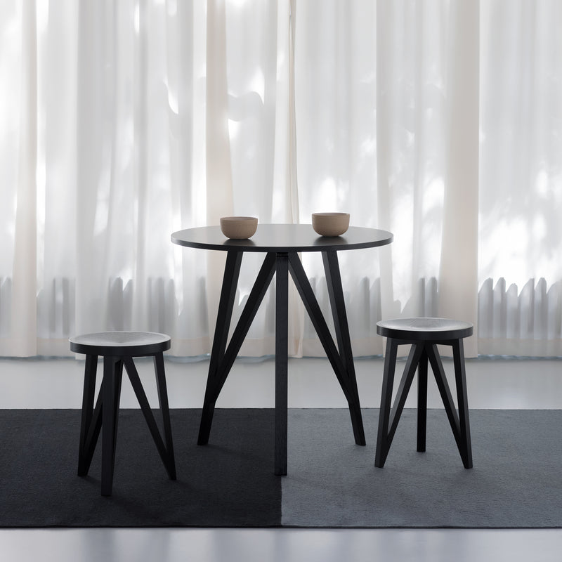 The JL3 Faber Bistro Table by Loehr is a minimalist dining set with a round black table and two matching stools on a two-toned rug. Made from solid oak, it captures northern European design. Two bowls adorn the table, while white curtains softly diffuse light in the background.