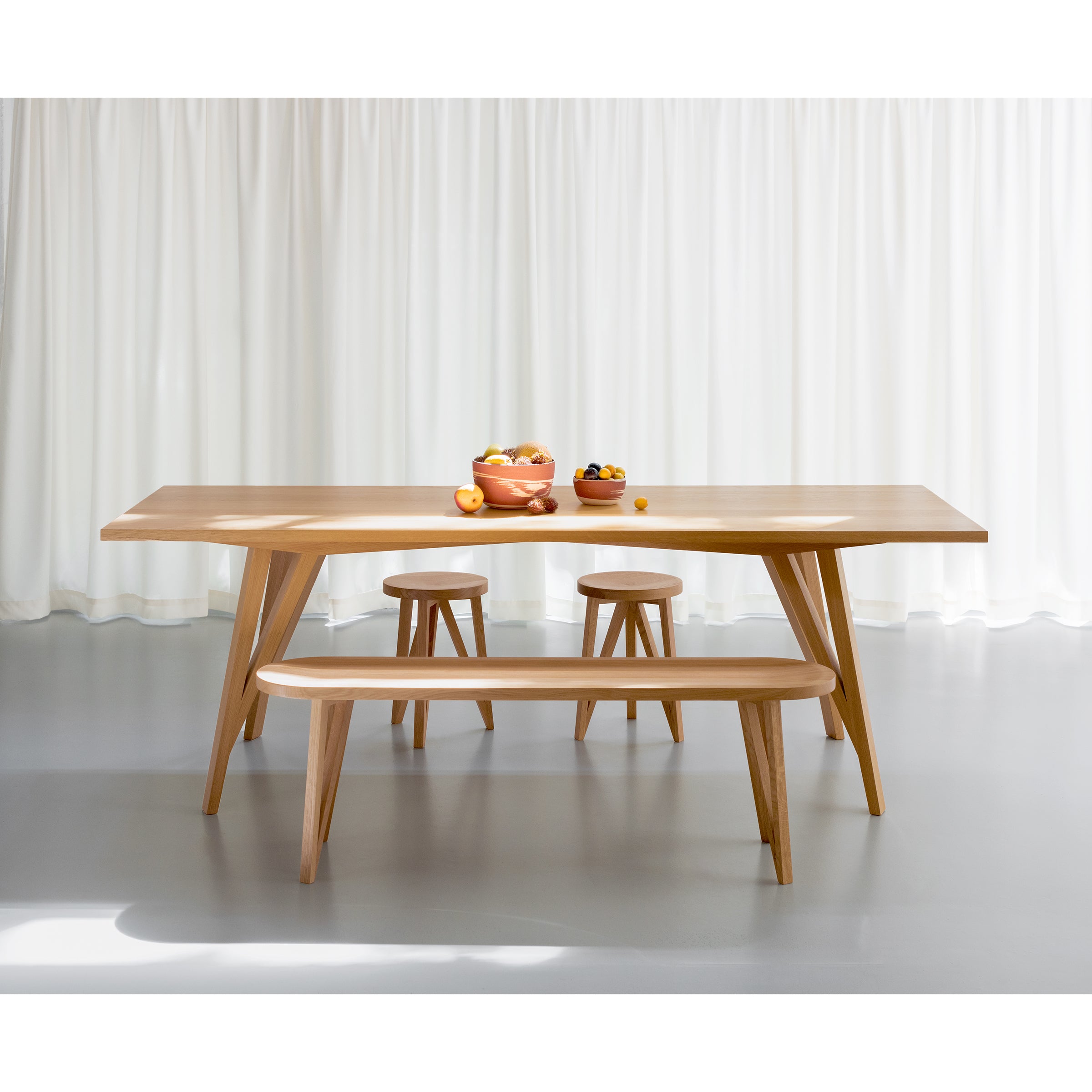 A minimalist dining setup showcases the Loehr JL5 Sabeth Table paired with a bench and three stools, adorned with two bowls of assorted fruits. White curtains and a smooth gray floor emphasize the craftsmanship of this simple, modern scene.