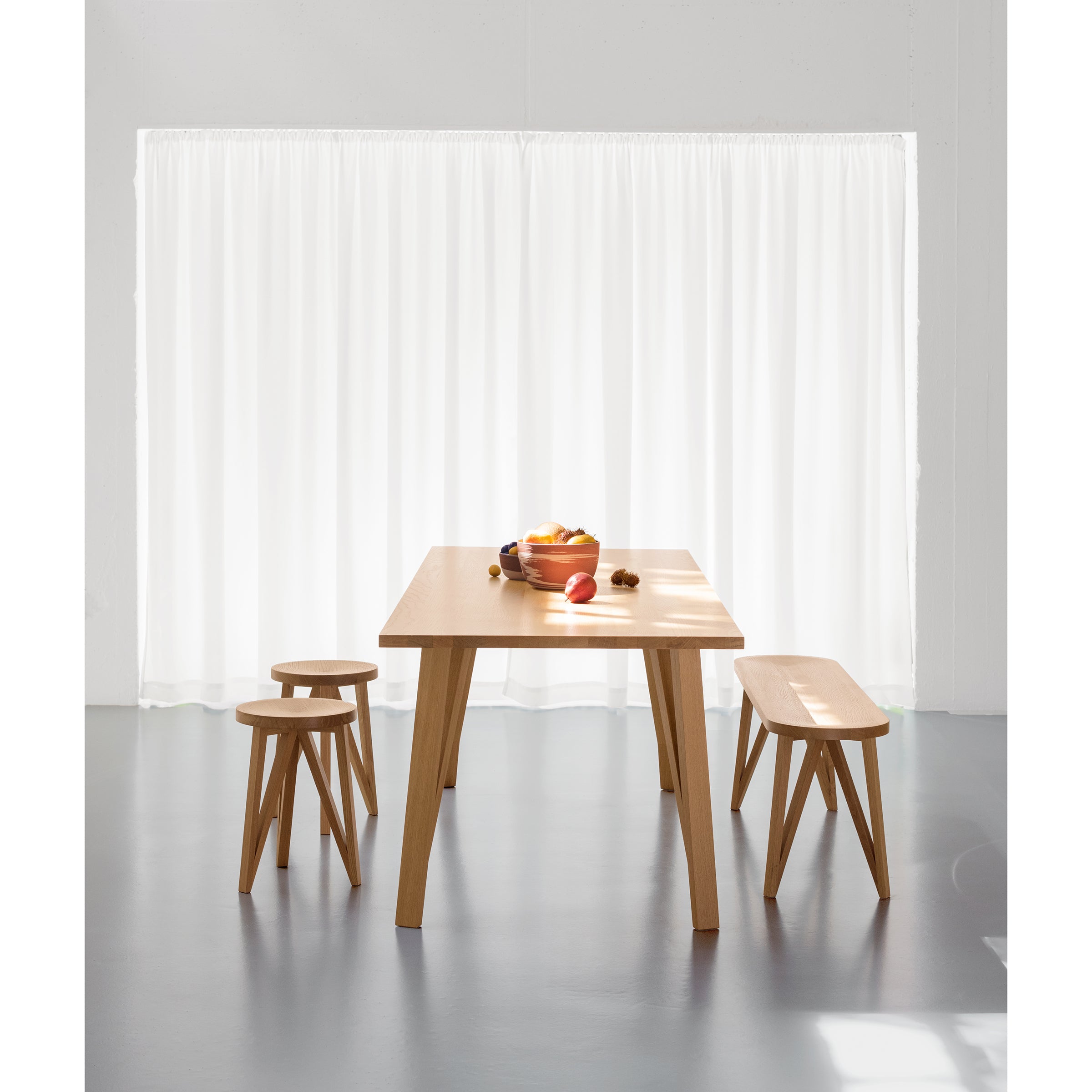 A minimalist dining setup highlights the Loehr JL5 Sabeth Table with a bench and two stools on a sleek gray floor. A fruit bowl with apples and bananas adorns the table, while sheer white curtains gracefully drape over a large window in the background.