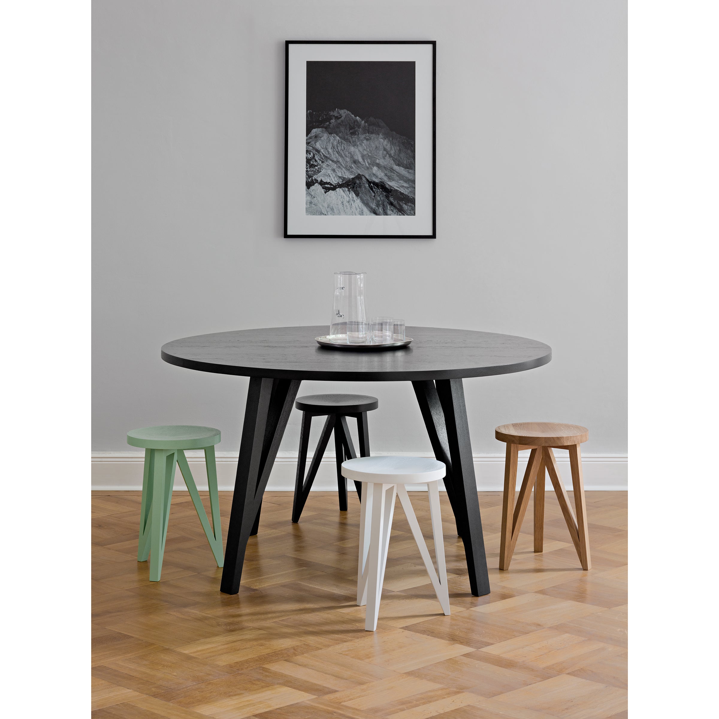 A JL6 Sabeth Round Table by Loehr, crafted impeccably, holds a glass carafe and trays. Its encircled by four wooden stools in green, white, black, and beige. A black-and-white mountain photo adorns the gray wall above a light wood herringbone-patterned floor.