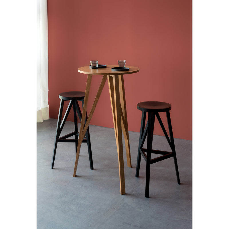 The JL7 Faber High Table by Loehr, a round wooden table crafted from solid oak, stands against a muted red wall alongside two matching black stools. It reflects northern European design and is bathed in soft natural light, highlighting the neatly placed plates and glasses.