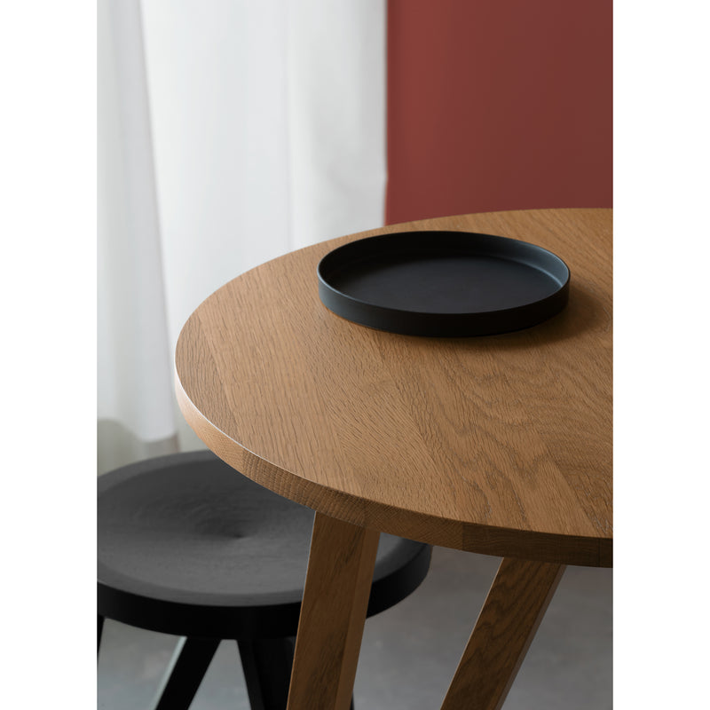 The JL7 Faber High Table by Loehr showcases northern European design with its round wooden build and black circular tray. Nearby is a black stool with a round seat, set against a sheer white curtain and red wall, offering an elegant contrast.