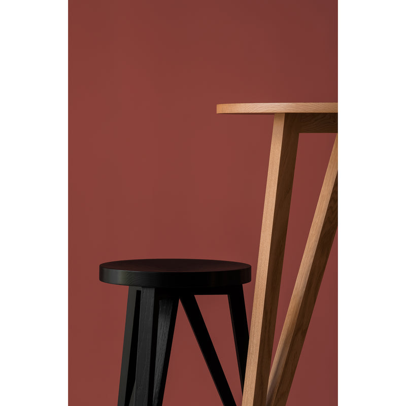 The JL7 Faber High Table by Loehr, with its solid oak and angled legs, pairs beautifully with a black stool featuring a round seat. Set against a reddish-brown background, this arrangement exudes northern European design charm with its geometric lines and contrasting colors.