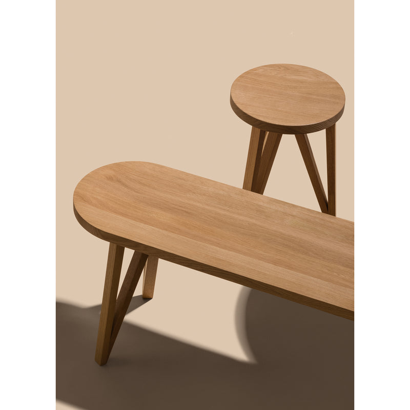 The JL8 Faber Bench and stool by Loehr, crafted in a minimalist northern European style, feature solid oak designs with an elongated oval bench and circular stool. Set against a neutral background, they have simple angled legs that cast soft shadows on the floor.