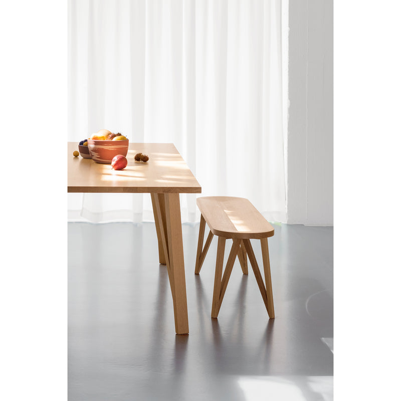 A JL8 Faber Bench by Loehr sits against light curtains with a table holding a wooden bowl of colorful fruit, including apples and oranges. The minimalist, solid oak design evokes a calm, modern northern European aesthetic.