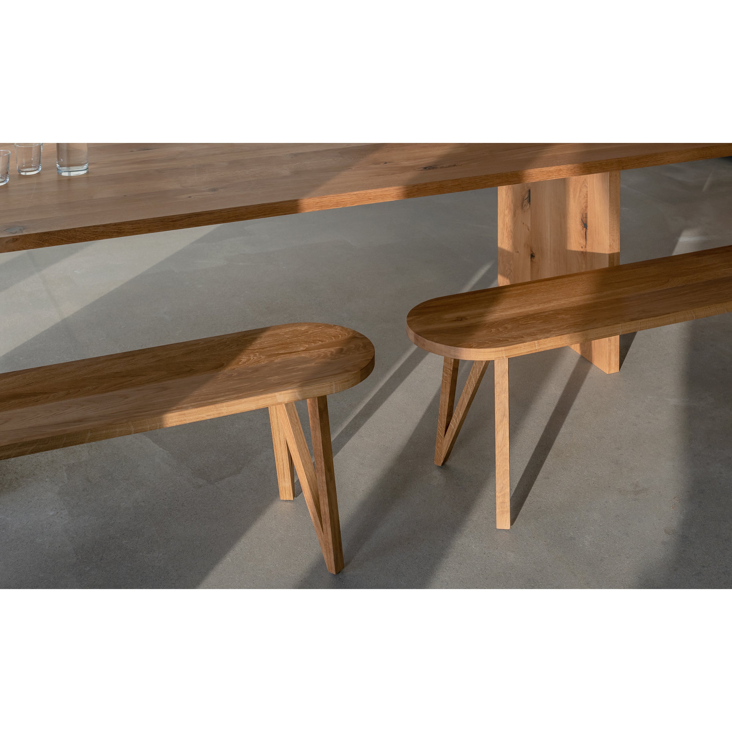 A JL8 Faber Bench from Loehr, with its northern European design, smooth edges, and natural wood grain, is bathed in sunlight on a concrete floor. A few glasses rest on the solid oak table, with shadows from the furniture adding depth to the scene.