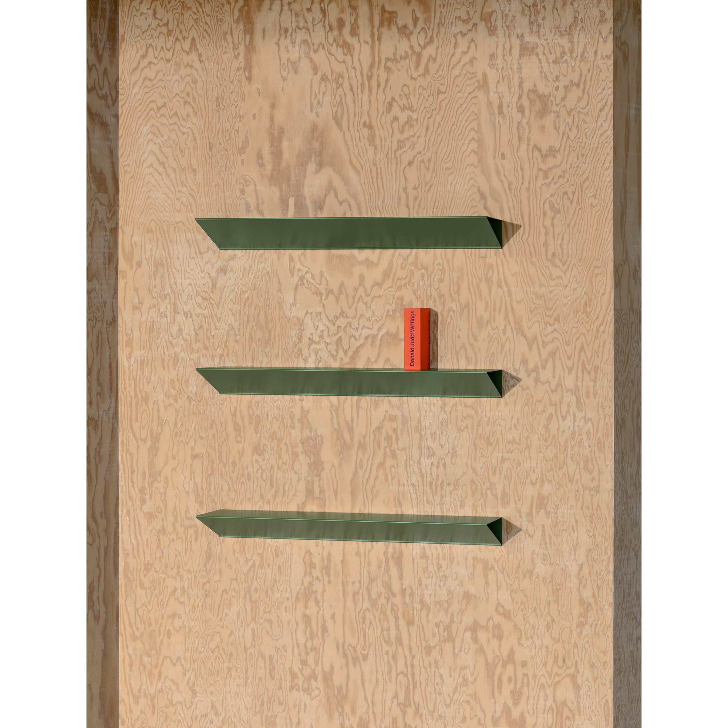 Three green Loehr L1 Newton shelves with a minimalist triangular profile are mounted on a wooden wall. The middle shelf, made of sheet steel, displays an upright red book.