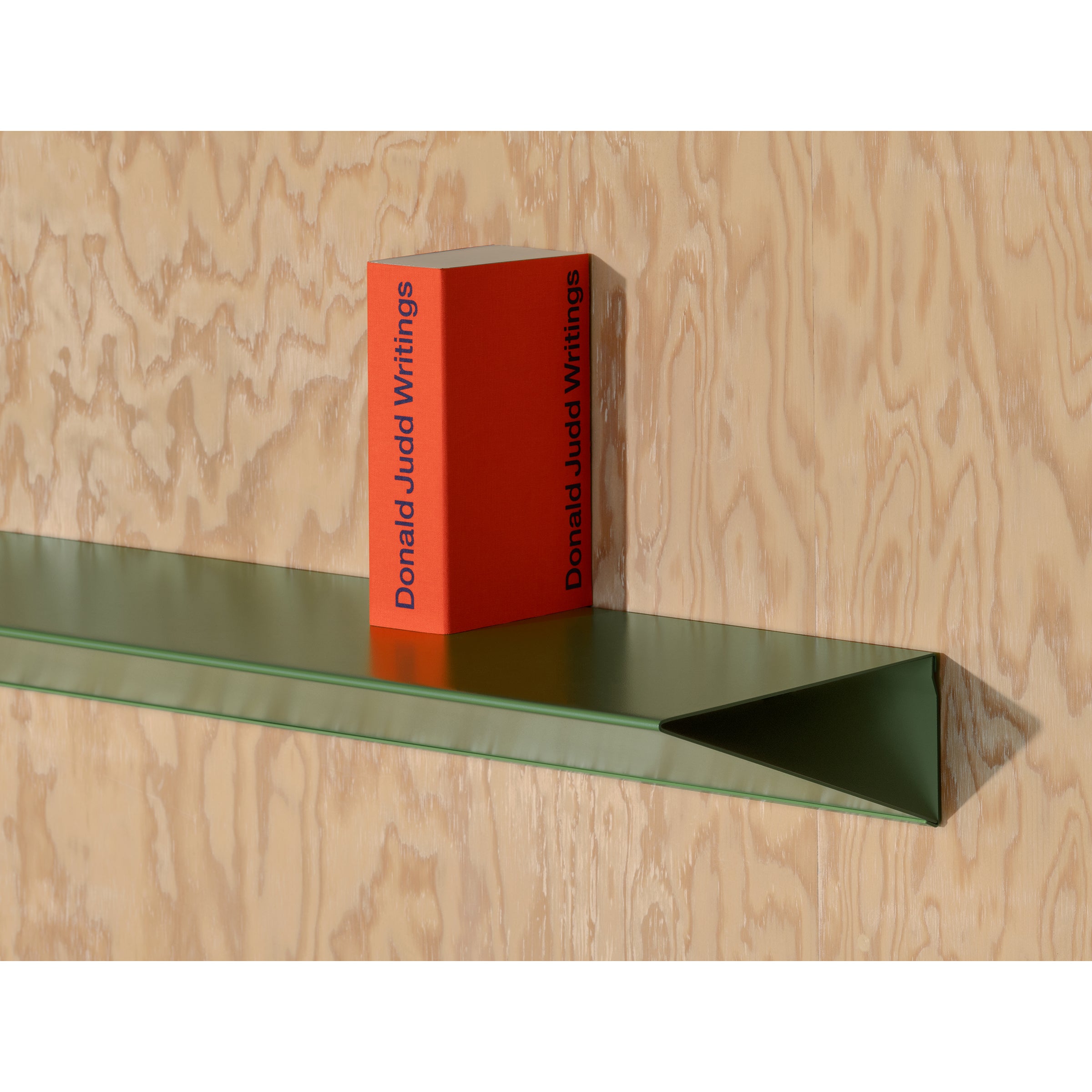 Donald Judd Writings, a red book, stands upright on Loehrs L1 Newton Shelf, boasting a minimalist triangular profile made from sleek sheet steel against a light wood-textured wall.