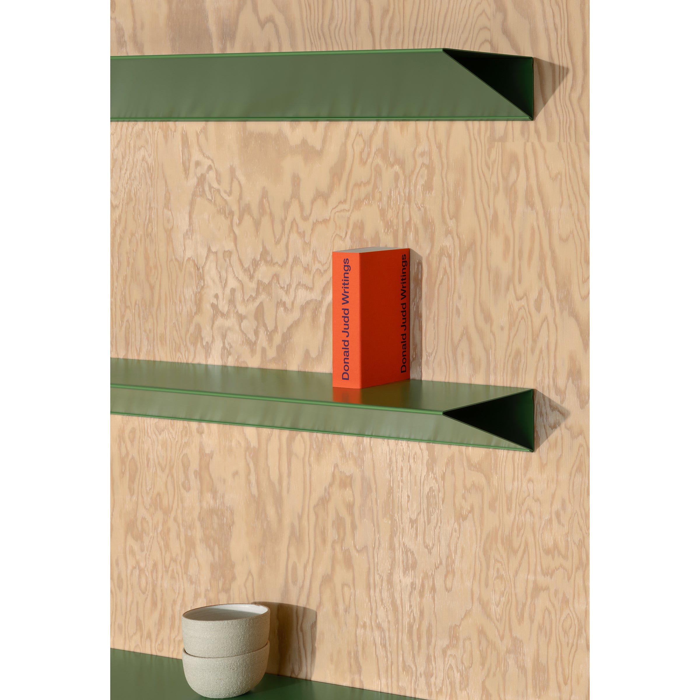 A minimalist wooden wall showcases three green shelves. The top shelf, an L1 Newton Shelf by Loehr, elegantly holds an orange book. Below is a small round bowl on the bottom shelf, while the unoccupied middle shelf maintains simplicity with its triangular profile.
