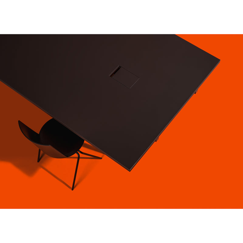 A minimalist scene showcases a Loehr L2 Eno Worktable with clean lines against an orange backdrop, with part of a black chair visible beneath the tables corner.