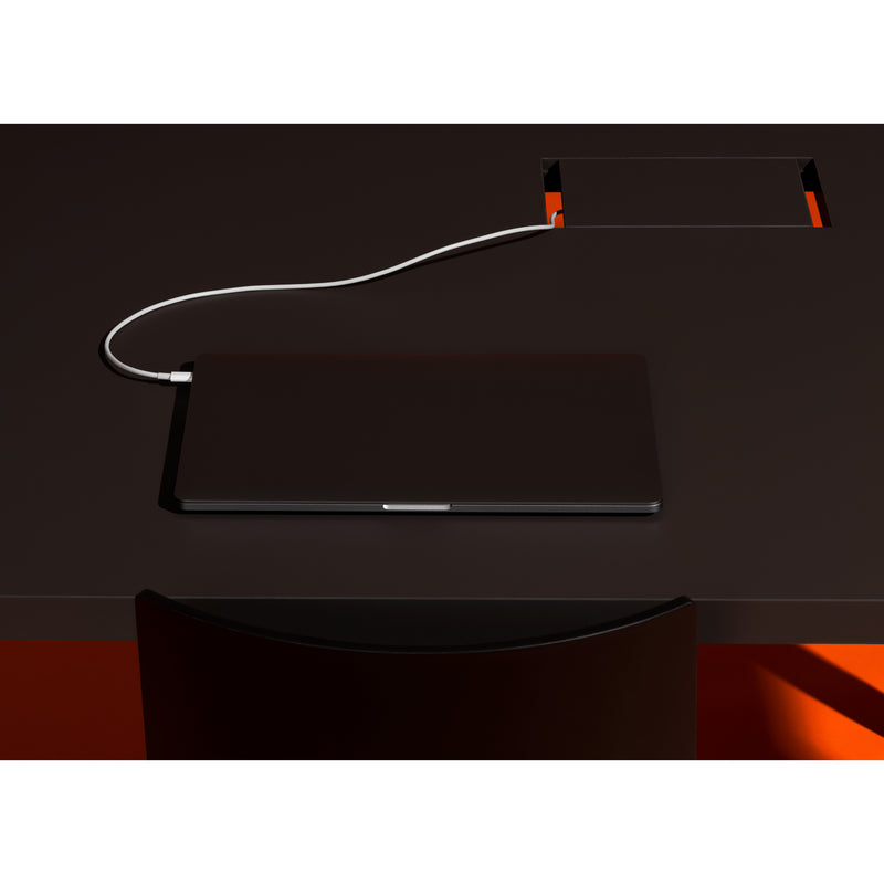 A closed laptop rests on a dark Loehr L2 Eno Worktable, plugged into a power outlet with a white cable. The desk features a rectangular cutout displaying an orange surface beneath. A black chair is partially seen in front of this sleek hybrid workstation.