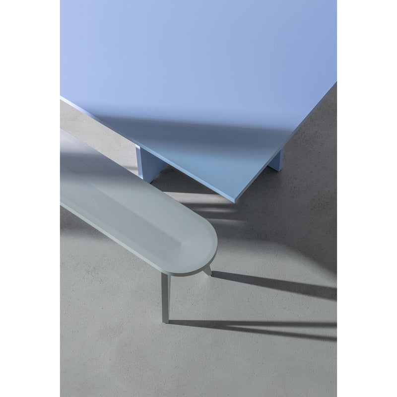 A minimalist scene showcases the corner of a Loehr L4 BASIL TABLE in sky-blue with a light gray bench on a smooth floor. The sculptural design and shadows from the coated fiberboard evoke a modern, serene vibe.