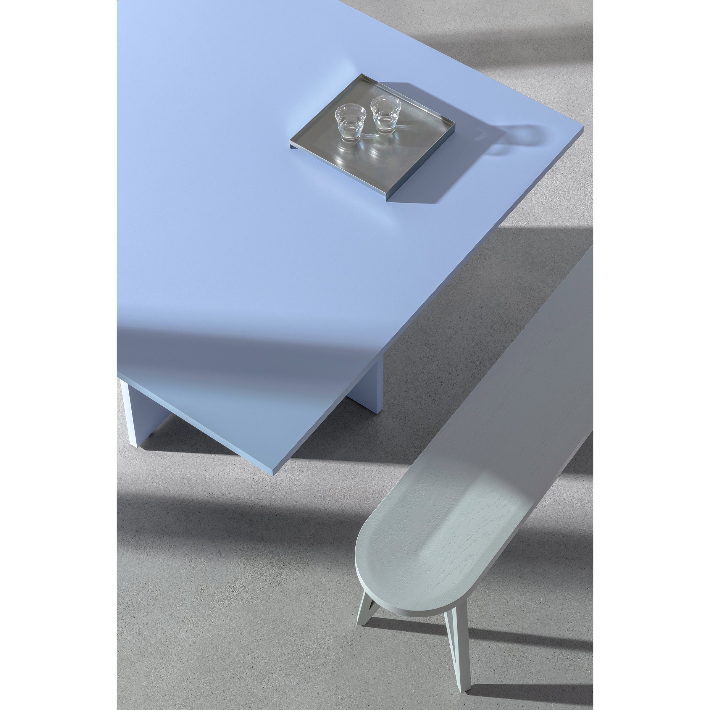 A minimalist scene showcases the L4 BASIL TABLE by Loehr with a sculptural design. A silver tray holds two glass cups, complemented by a pale gray bench nearby. Both cast soft shadows on the smooth light gray floor, creating elegant harmony under gentle lighting.