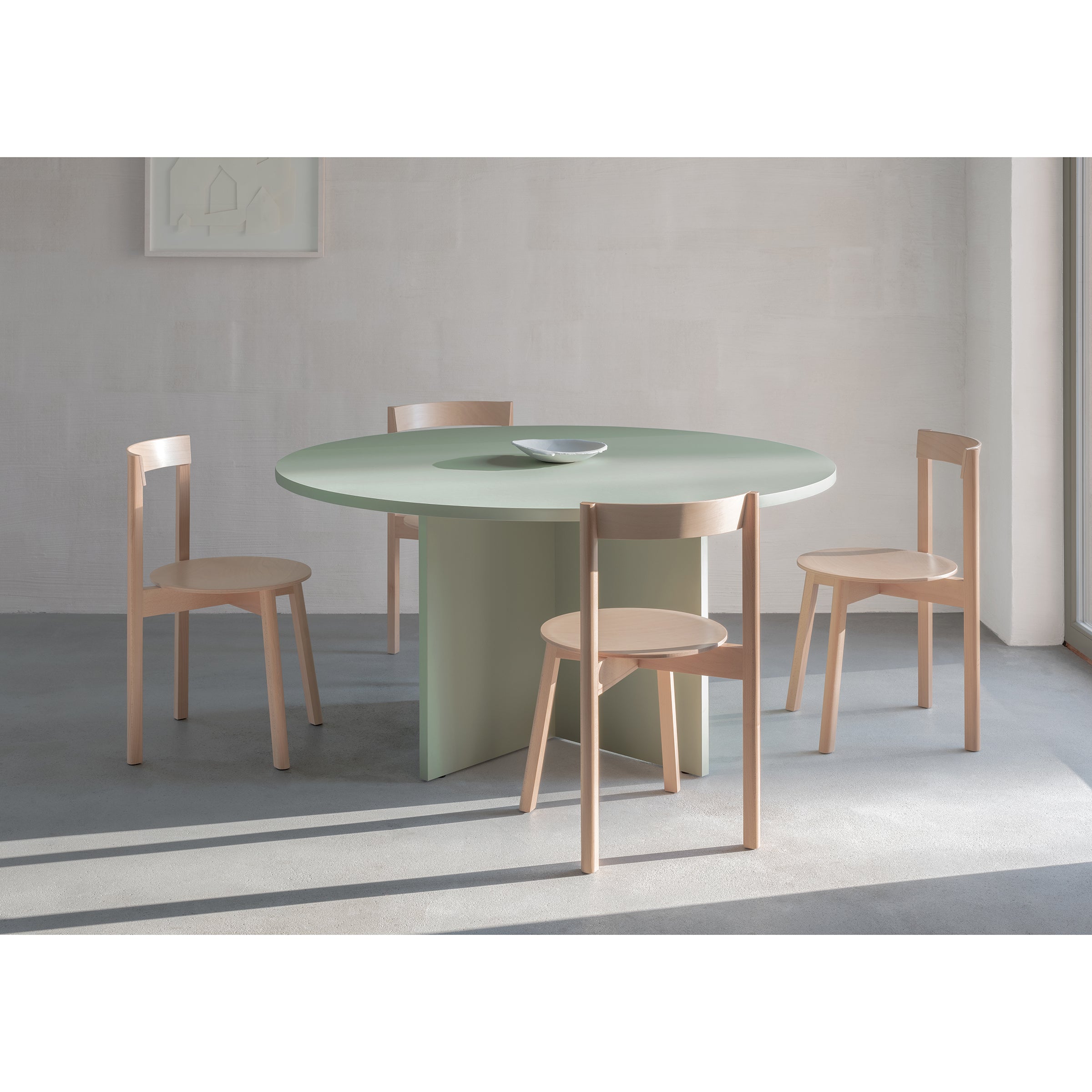 A minimalist dining room features a round mint-green L4 BASIL TABLE by Loehr, with a central pedestal base and four light wooden chairs. Natural light streams in, casting subtle shadows and enhancing its sculptural look with a small dish at the center.