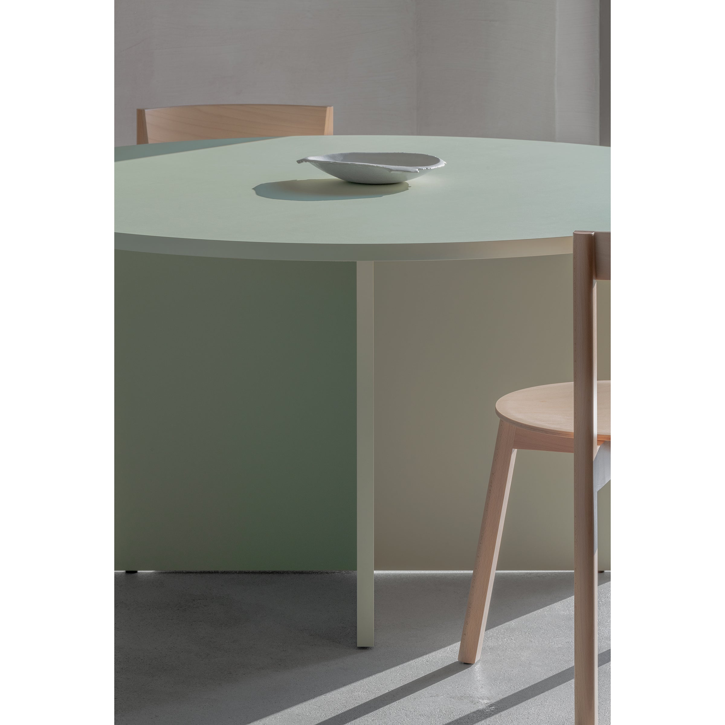 The Loehr L4 BASIL TABLE, with its sculptural design, showcases a light green round surface made from coated fiberboard. Complemented by two light wood chairs and a small ceramic bowl, this minimalist setup resides in a softly lit room with neutral walls.