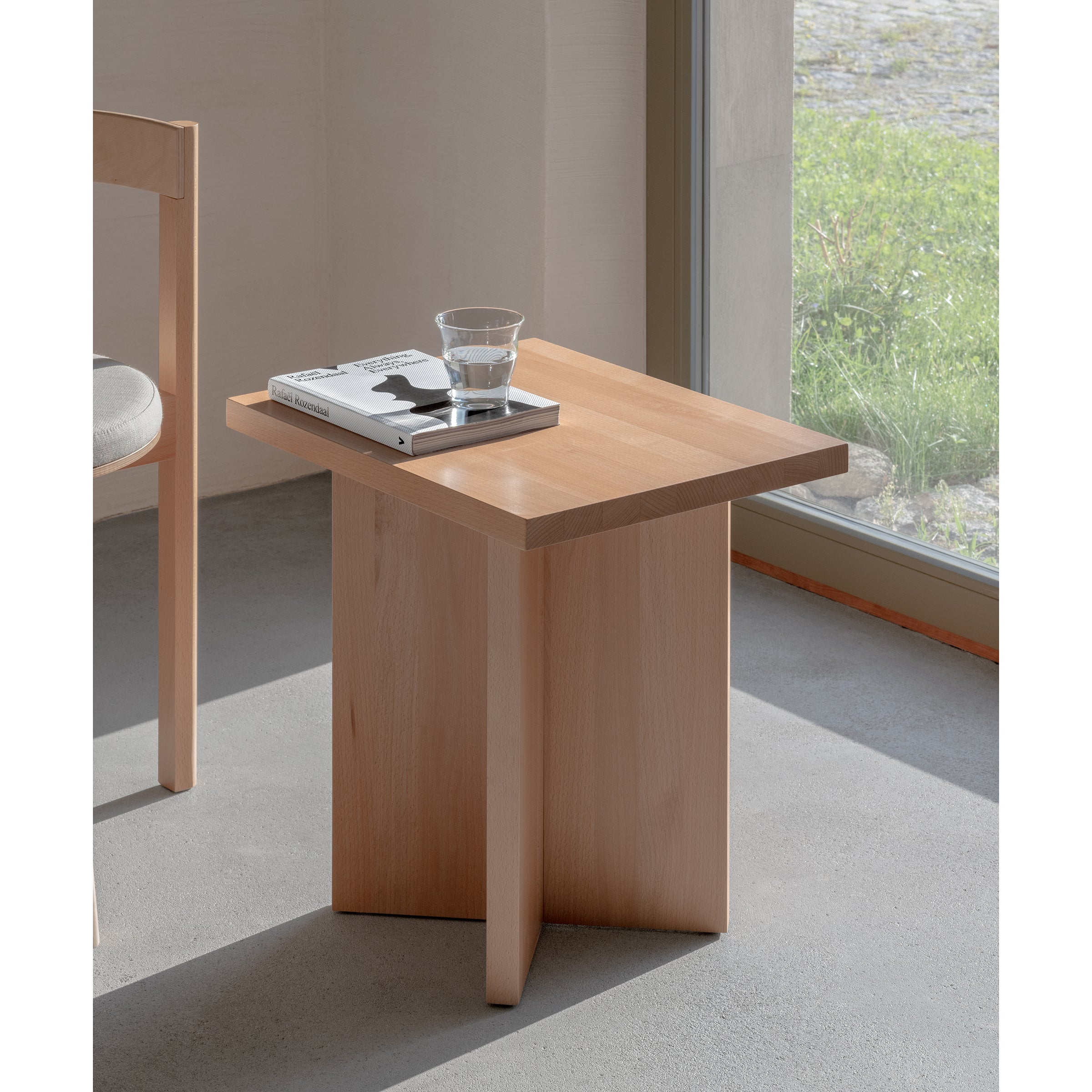 The L6 Corinth Side Table by Loehr, made of solid beech and featuring a geometric design, elegantly rests by a window. It supports a glass of water and a closed book. Nearby, part of a chair with a light-colored cushion is visible as sunlight filters in, casting gentle shadows on the floor.