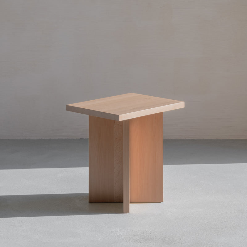 The L6 Corinth Side Table by Loehr is a sculptural piece crafted from solid beech with a minimalist rectangular top and intersecting vertical base, placed on a smooth light gray floor next to a pale wall.