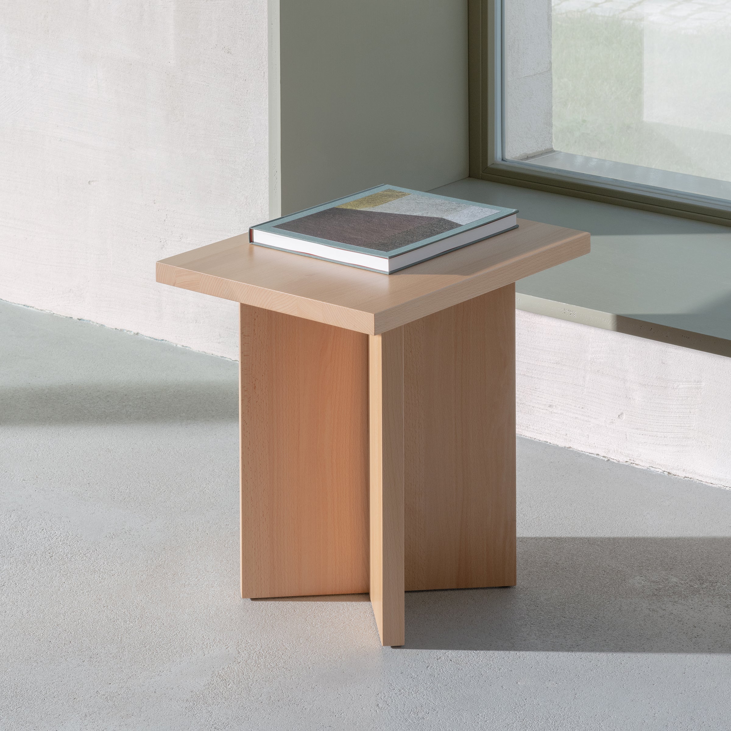 The minimalist L6 Corinth Side Table by Loehr is in a bright room near a window. A gray-covered book sits atop this solid beech piece, harmonizing with the concrete floor and light-colored walls, enhancing its sculptural appeal.