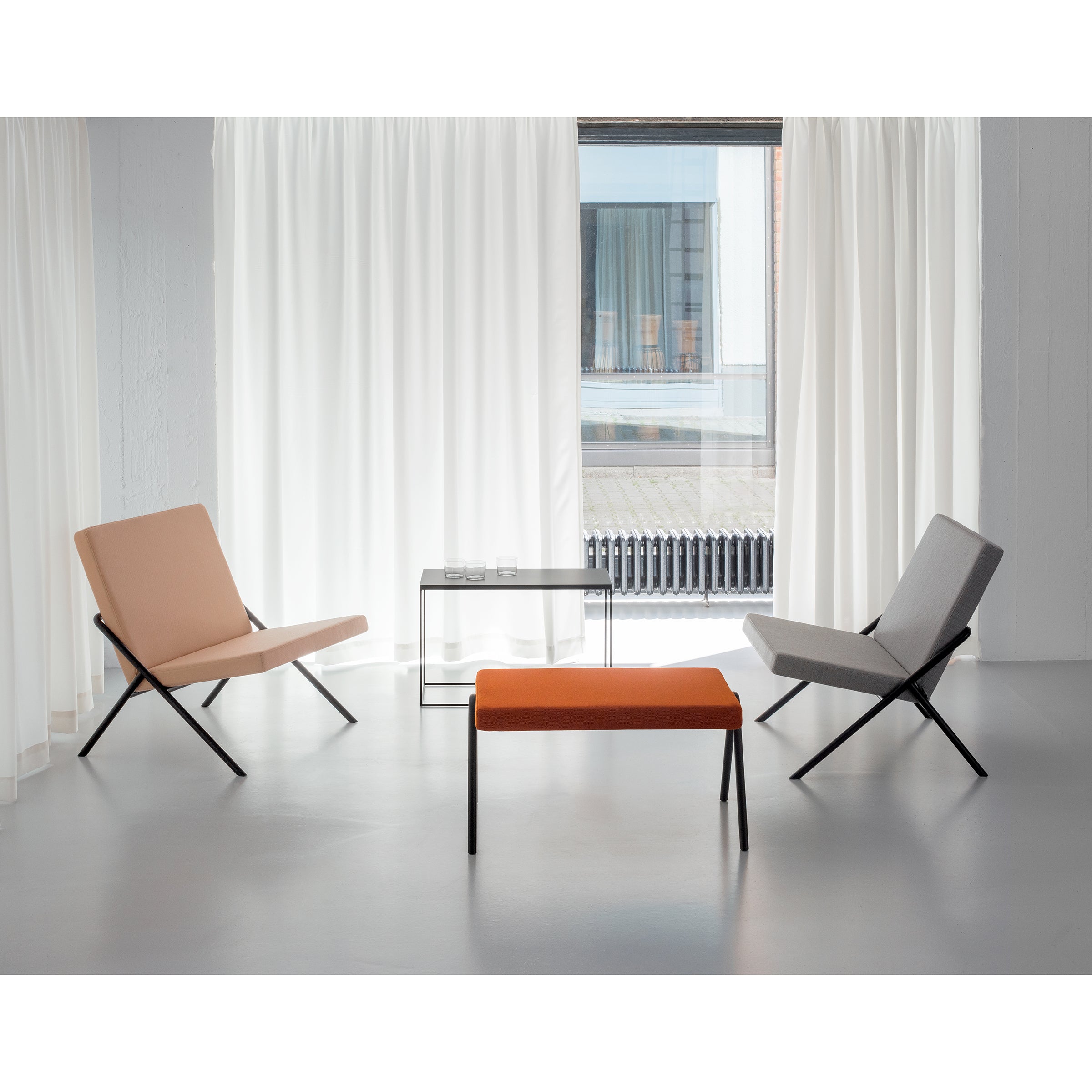 A minimalist room features a beige chair, a gray chair, and the DL6 Plato Ottoman by Loehr in orange with luxurious seating. A glass coffee table holds two glasses beside sheer white curtains over a large window showcasing urban scenery.