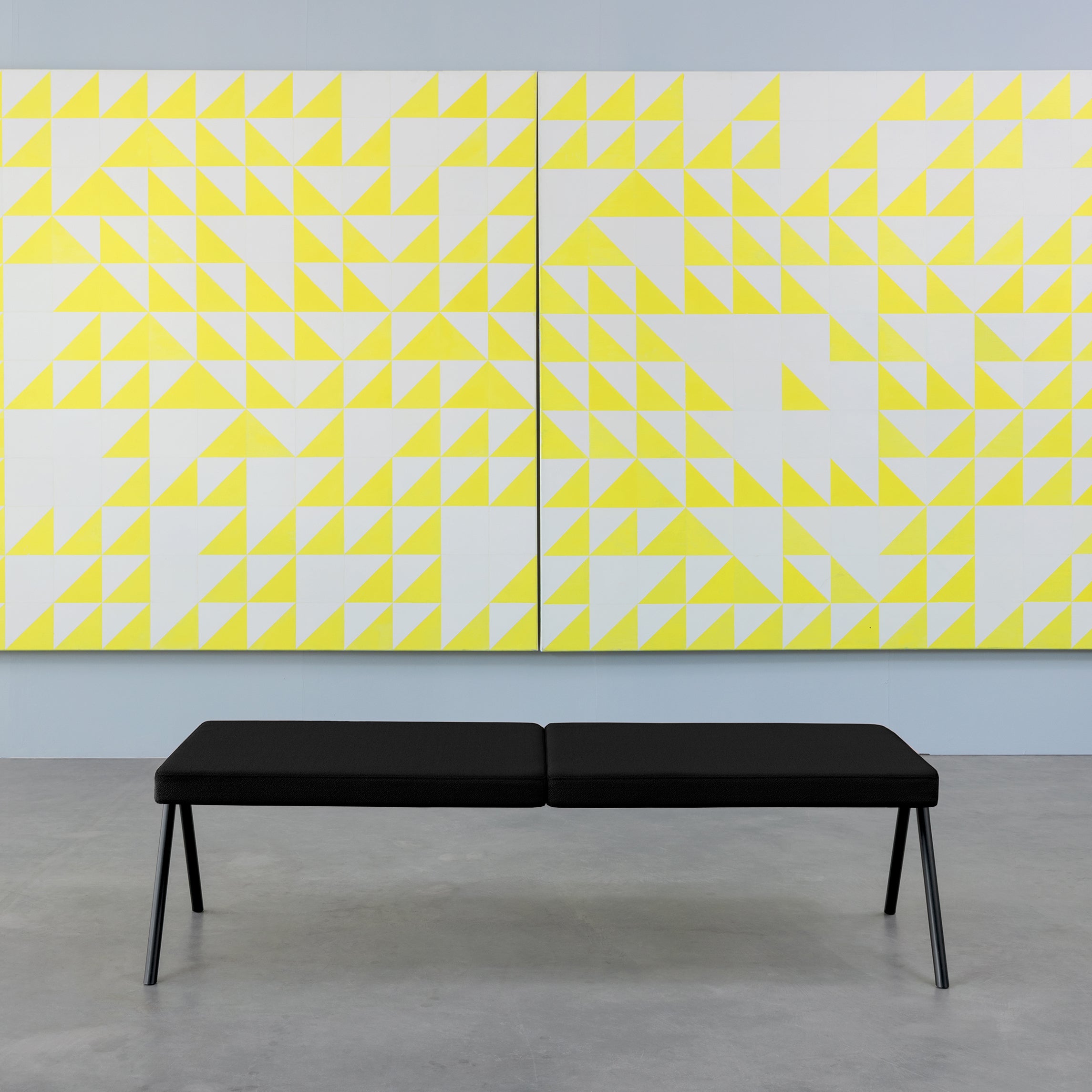 The DL6 Plato Bench by Loehr, in an elegant black finish, rests on a sleek gray floor beneath a large, abstract artwork featuring yellow and white geometric triangles against a light blue wall.