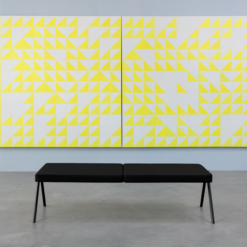 The DL6 Plato Bench by Loehr, in an elegant black finish, rests on a sleek gray floor beneath a large, abstract artwork featuring yellow and white geometric triangles against a light blue wall.