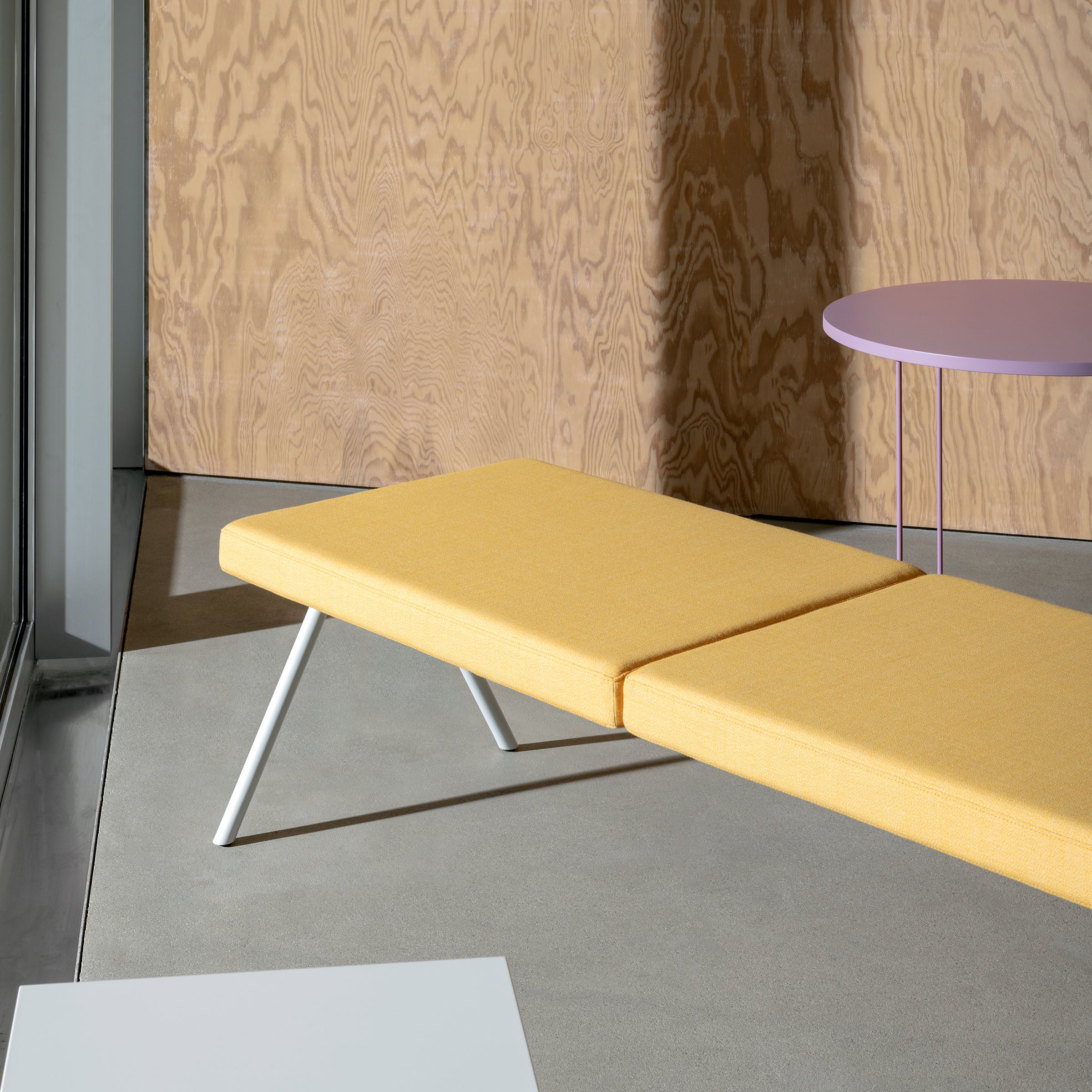 A modern interior features the DL6 Plato Bench by Loehr, upholstered in vibrant yellow, paired with a round lavender table and wooden wall paneling. Natural light through a large window bathes this elegant minimalist arrangement.