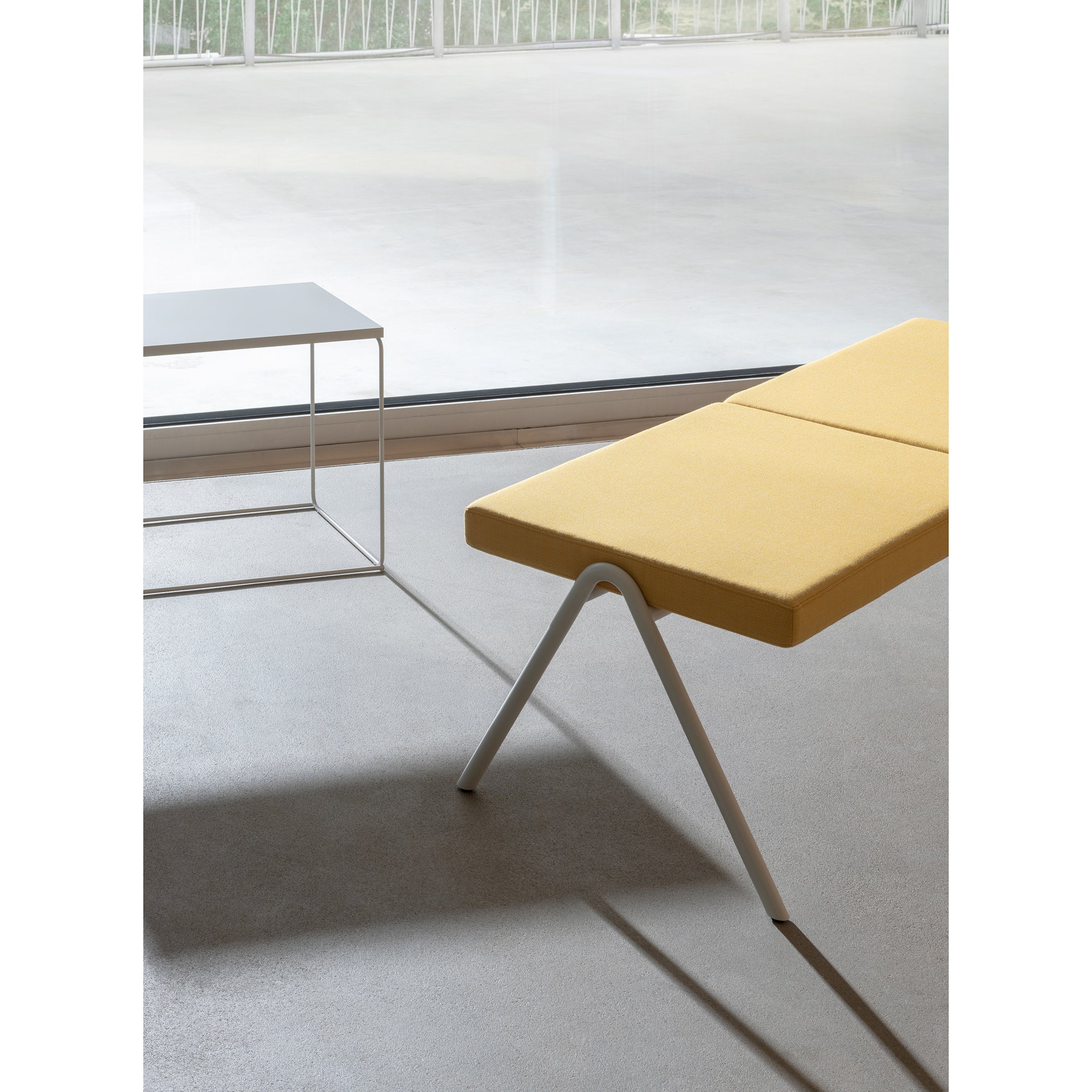 A minimalist interior features the DL6 Plato Bench by Loehr, with an upholstered yellow seat and angled white base, beside a simple white square table. Daylight pours in through a large window, creating soft shadows across the light gray floor.
