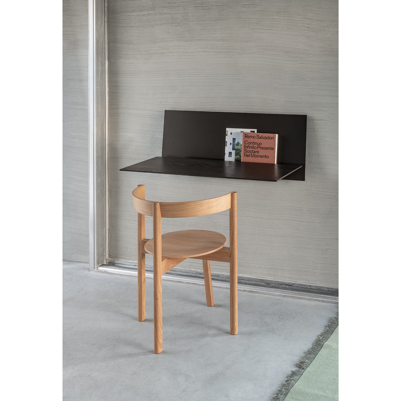 A minimalist workspace showcases a Loehr S1 CHAPTER Bureau, featuring a floating black desk with a wooden chair. Books rest on the fold-up tabletop. The room has a concrete floor, subtly textured walls, and a green rug at the edge.