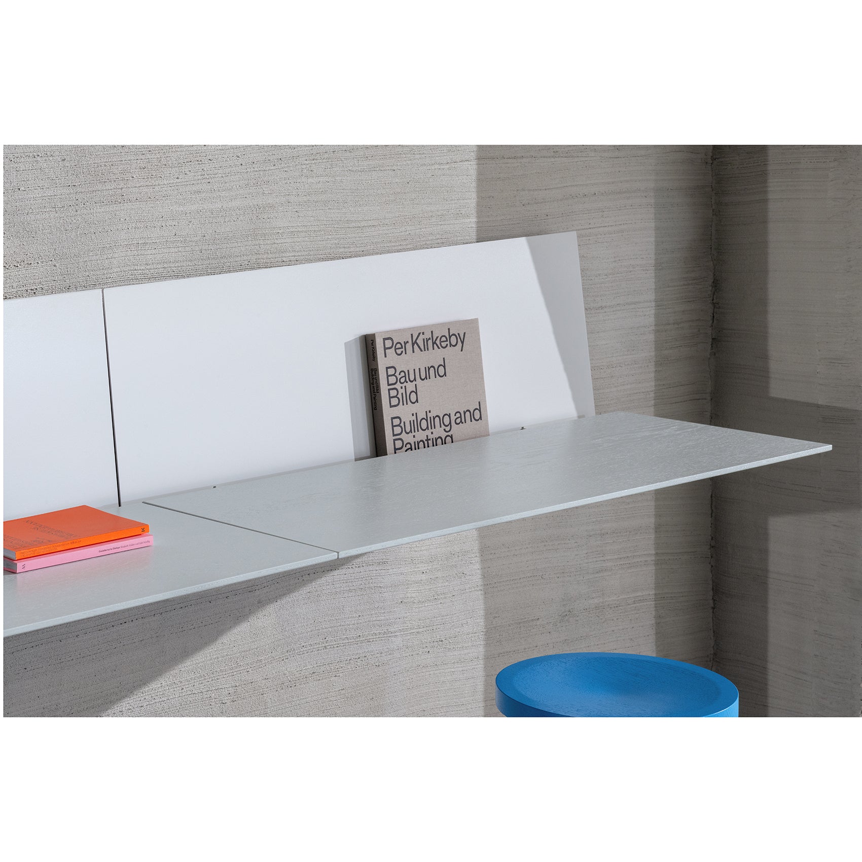 The S1 CHAPTER Bureau by Loehr is a minimalist wall-mounted desk with a gray finish, featuring the book Per Kirkeby: Bau und Bild and an orange notebook. A blue cylindrical stool sits below this stylish fold-up tabletop design, maximizing space efficiently.