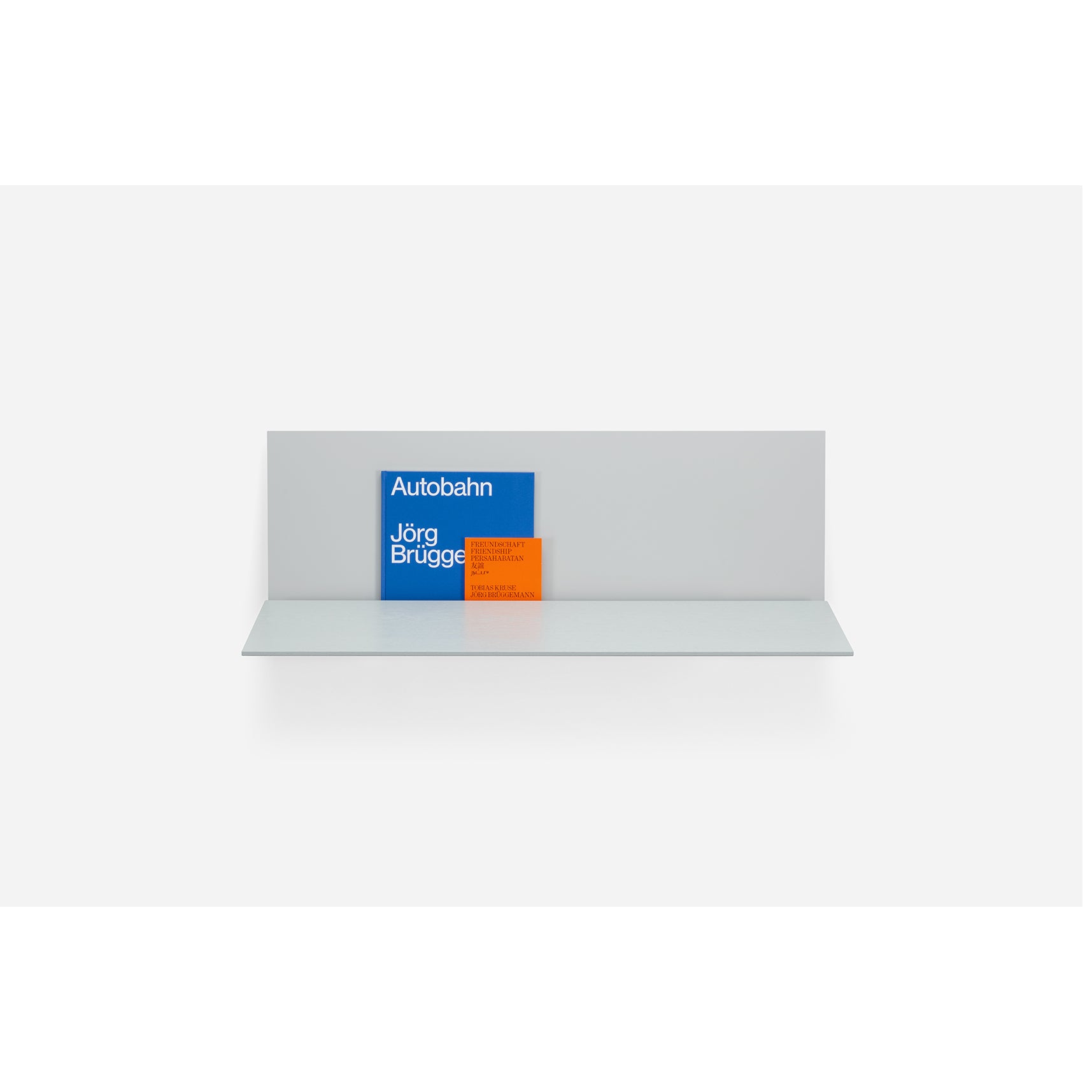 The S1 CHAPTER Bureau by Loehr is a minimalist wall-mounted shelf featuring a gray back panel that elegantly displays two books—Autobahn by Jörg Brügg as a larger blue book and another smaller orange one, creating a sleek, sophisticated setup against a white backdrop.