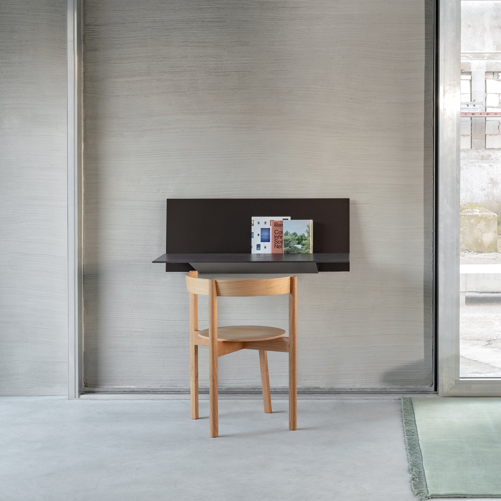 A minimalist workspace showcases the sleek black S1 CHAPTER Bureau by Loehr on a textured gray wall, with three books placed upright. A simple round-seated wooden chair is in front of the desk, and a plush rug is set on the concrete floor.