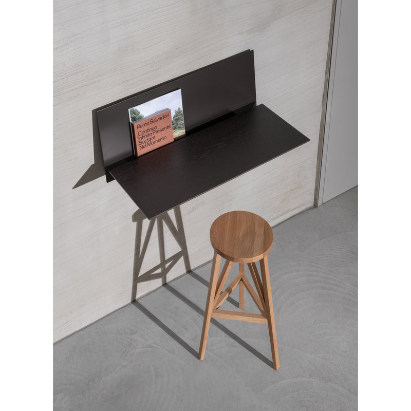 The minimalist setup includes a Loehr S1 CHAPTER Bureau, with a wooden stool placed before its sleek, satin black design against a light gray wall. A partially visible book lies on the bureaus shelf, casting shadows on the wall and floor.