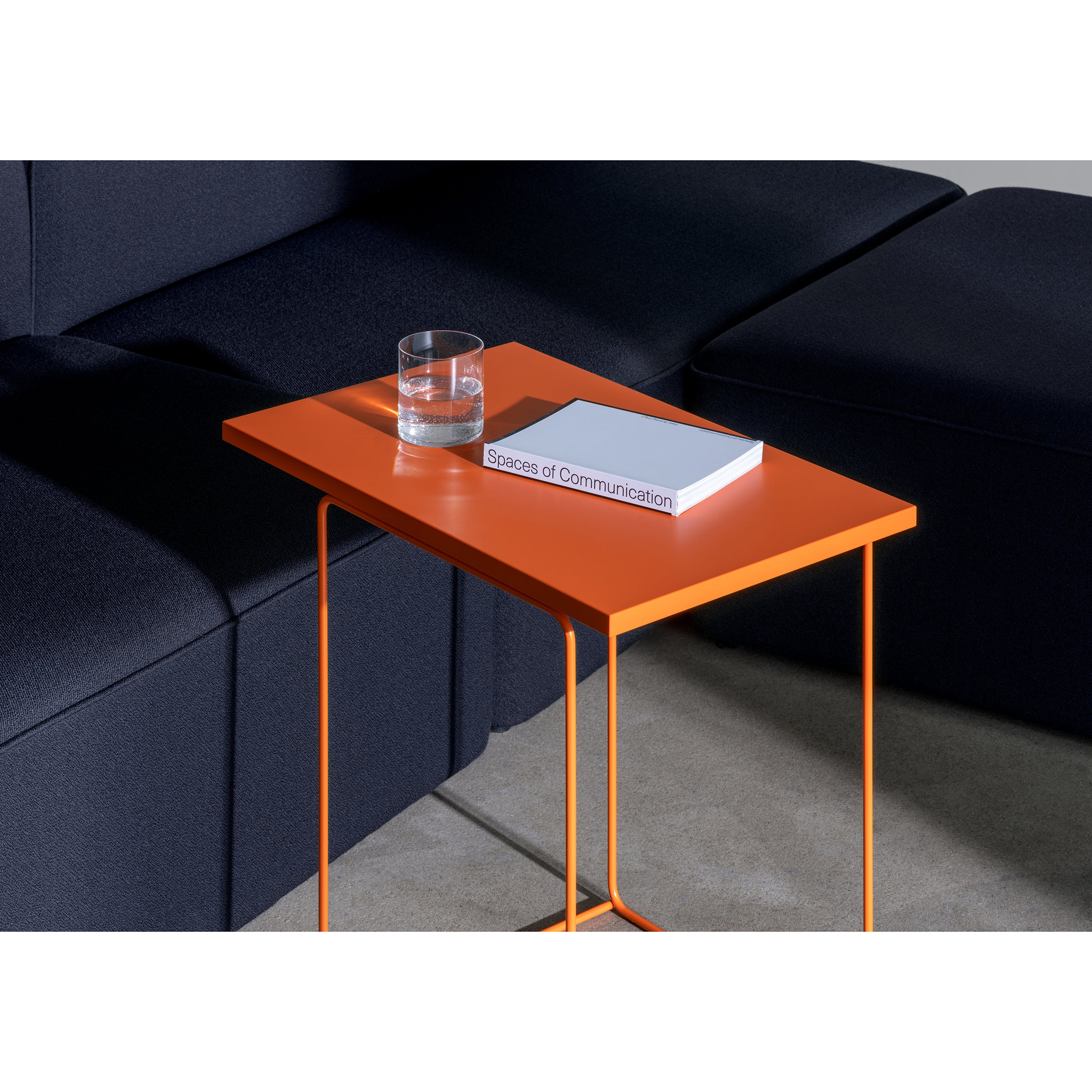 In a modern, minimalist living room, the DL1 Tangram Side Table Square by Loehr features an orange design next to a dark couch. The table holds a clear glass of water and Spaces of Communication, while a beige carpet elegantly complements the architectural landscape-inspired setup.