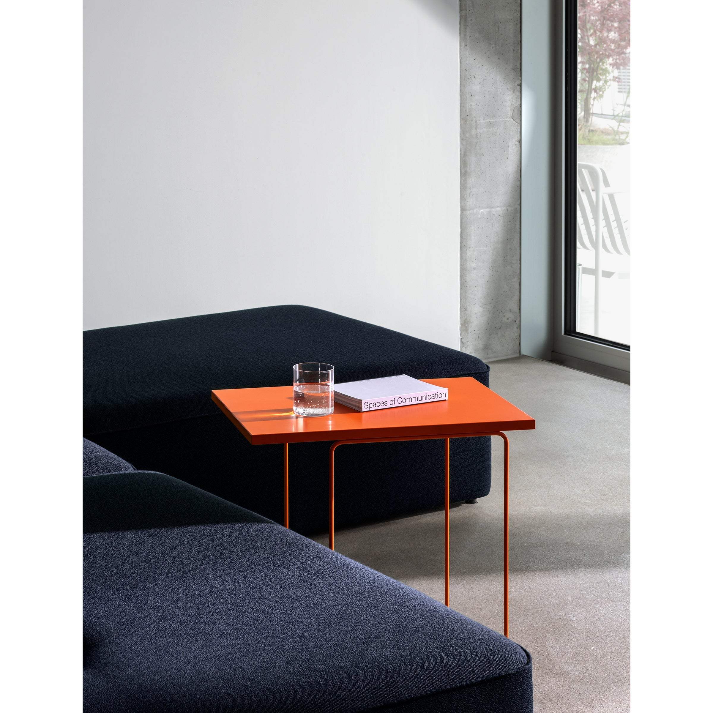 A minimalist room features a DL1 Tangram Side Table Asymmetric by Loehr, holding a glass of water and the book Spaces of Communication. Positioned between dark blue couches, it contrasts elegantly with a light gray wall and a large window.
