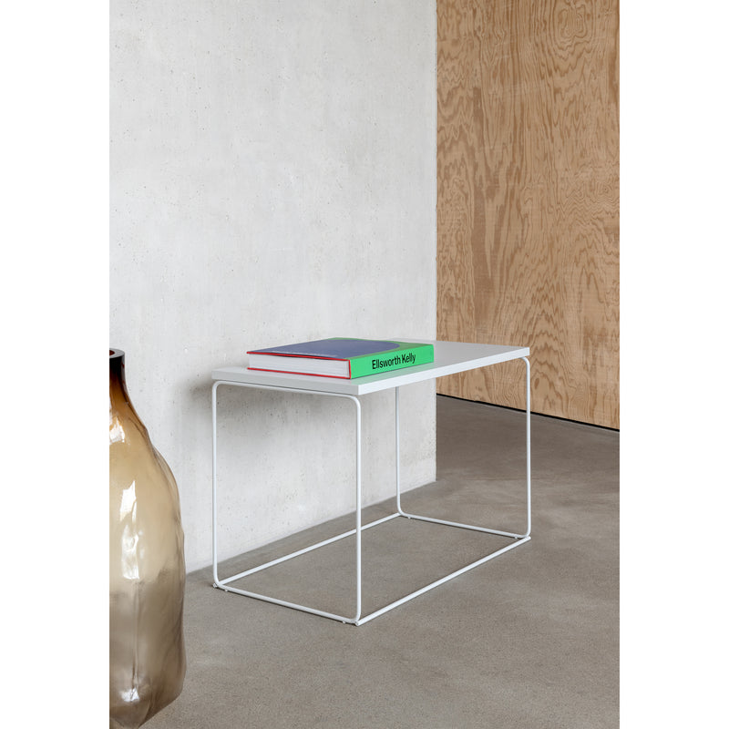A minimalist DL1 Tangram Side Table Rectangular by Loehr, crafted in white metal with a green book on top, stands against a plain concrete wall. A large beige ceramic vase sits to the left, complemented by the concrete floor and wooden panel for a serene architectural landscape.