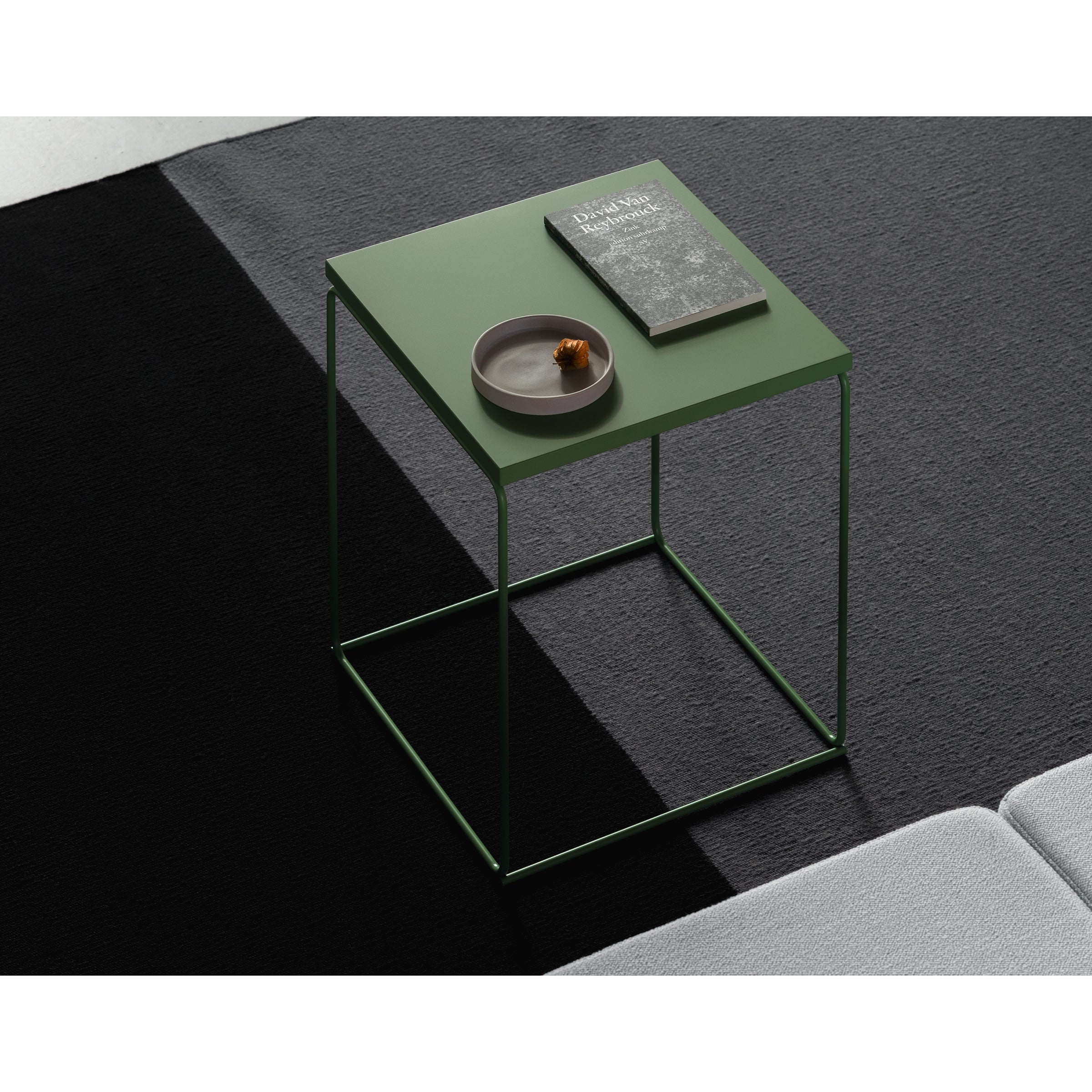 The minimalist DL1 Tangram Side Table Square by Loehr has a green top and thin legs, set on a black and white carpet. Its adorned with a brown snack dish and a dark book, beside the edge of a light gray sofa.