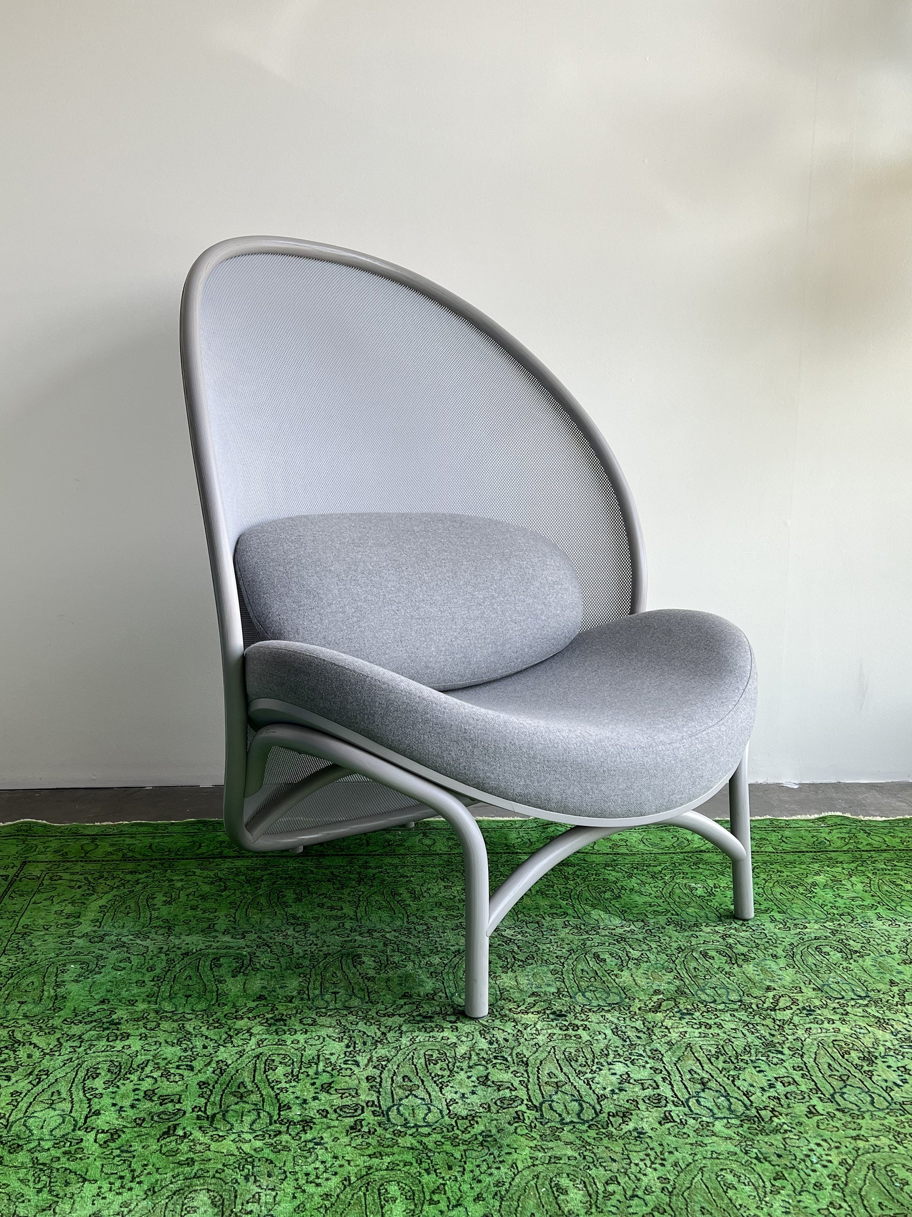 A gray Chips Lounge Chair by TON, featuring a modern curved backrest and comfortable upholstery, sits on a bright green patterned rug. Its minimalist design with smooth edges is elegantly displayed against a plain white wall.