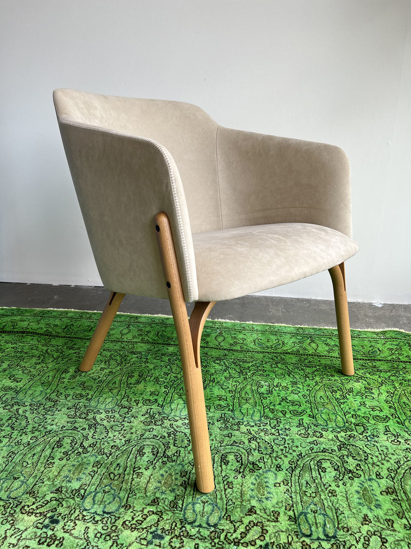 A beige, modern TON Lounge Chair Split with ergonomic upholstery and wooden legs rests on a vivid green patterned rug against a plain white wall, highlighting its finesse.