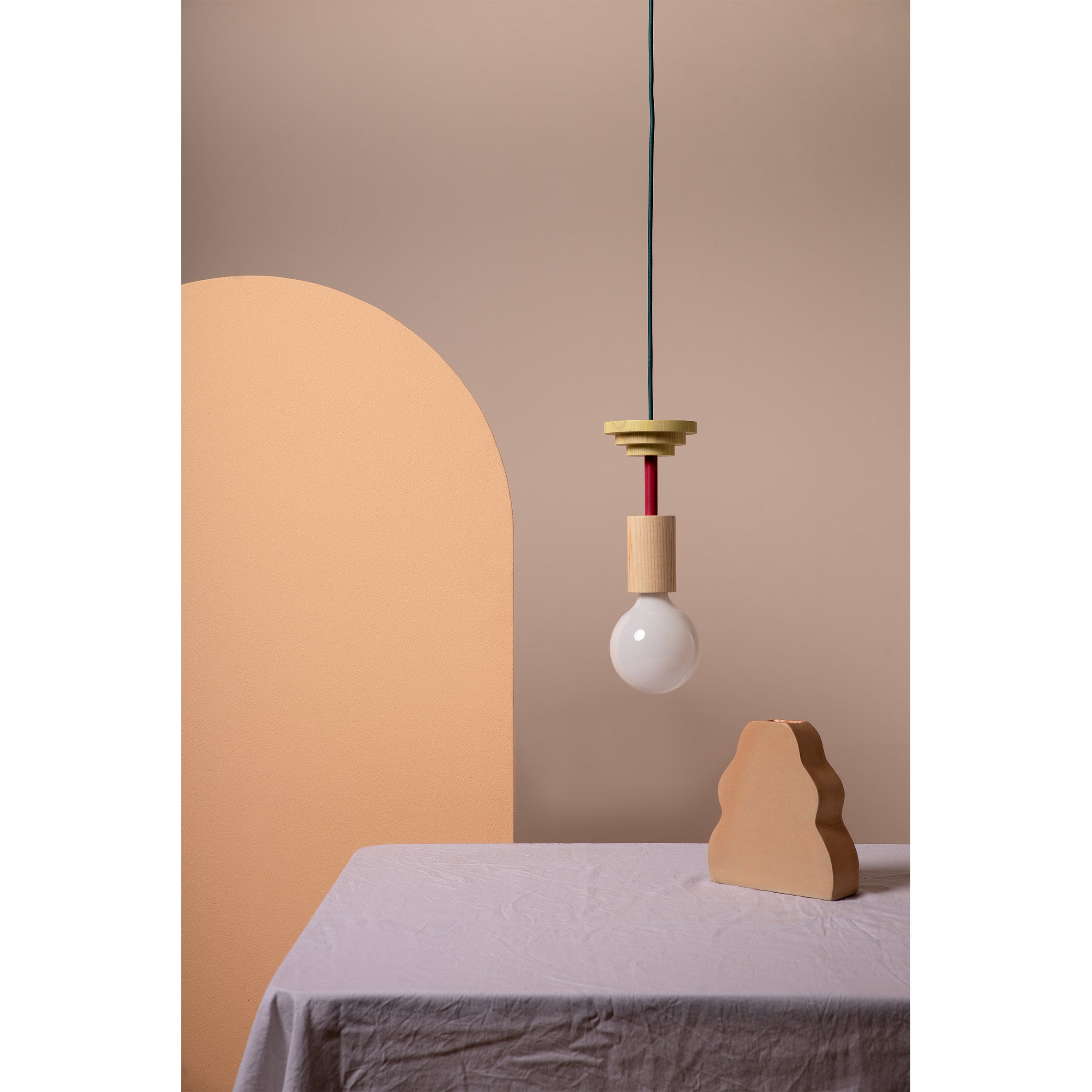 A minimalist room showcases Schneids Junit Mentis pendant lamp with a round bulb over a table with a white cloth. A curved, peach partition and an abstract clay sculpture add artistic flair to the beige background, enhanced by warm ash wood accents.