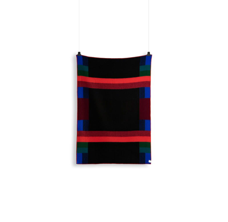 The Mikkel Large Throw by Roros is a colorful Norwegian wool tapestry with a Bauhaus-inspired geometric pattern of black, red, blue, and green stripes on a white background. Designed by Kristine Five Melvær, it features intersecting lines forming a rectangular shape.