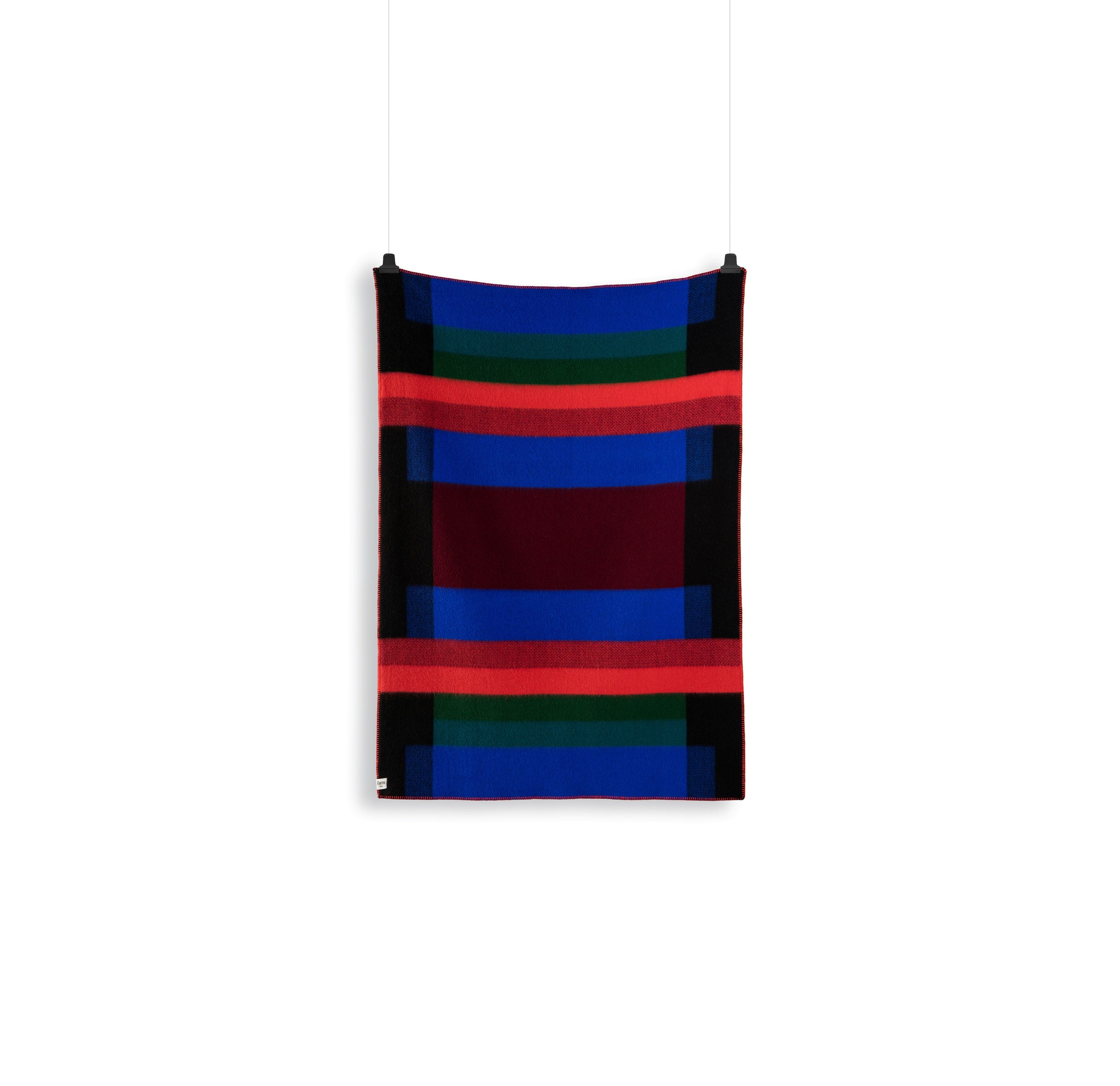 The Mikkel Large Throw by Roros displays a Bauhaus-inspired abstract geometric pattern with red, blue, green, and black rectangles and stripes. Crafted from Norwegian wool, it hangs on a white background with two clips at the top corners.