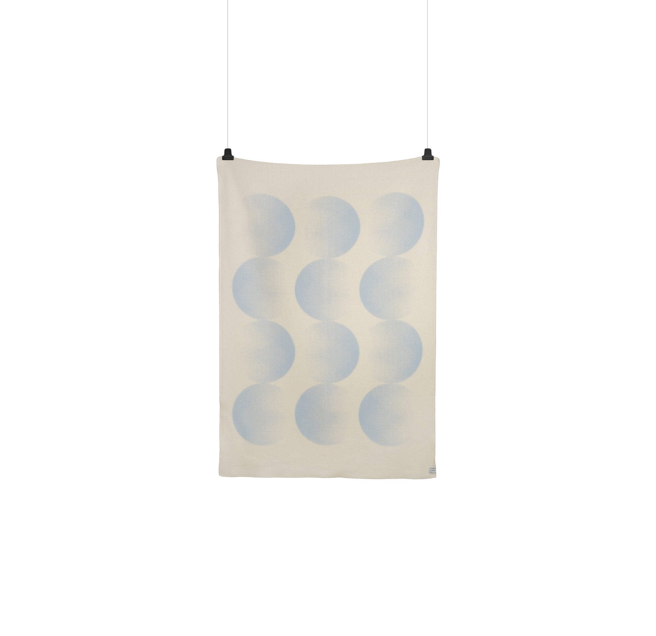 The Moon Large Throw by Roros, designed by Clara von Zweigbergk, is a hanging rectangular textile made from Norwegian lambswool. It features overlapping circular shapes in a gradient of light blue tones on a cream background and showcases a minimalist design.